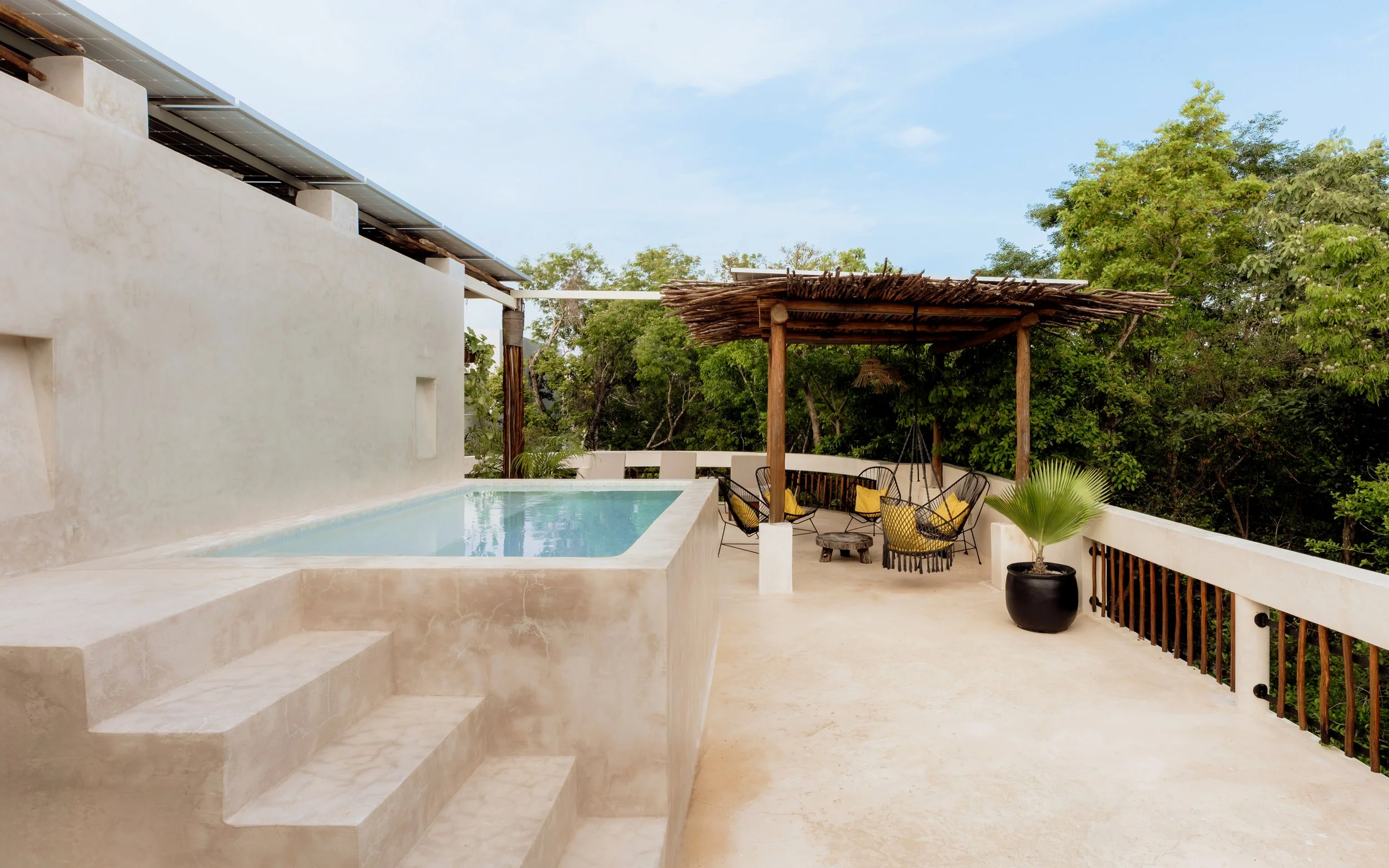 Outdoor patio with a small pool, seating area with black chairs and yellow cushions, a thatched roof structure, a potted plant, and surrounded by green trees under a blue sky.