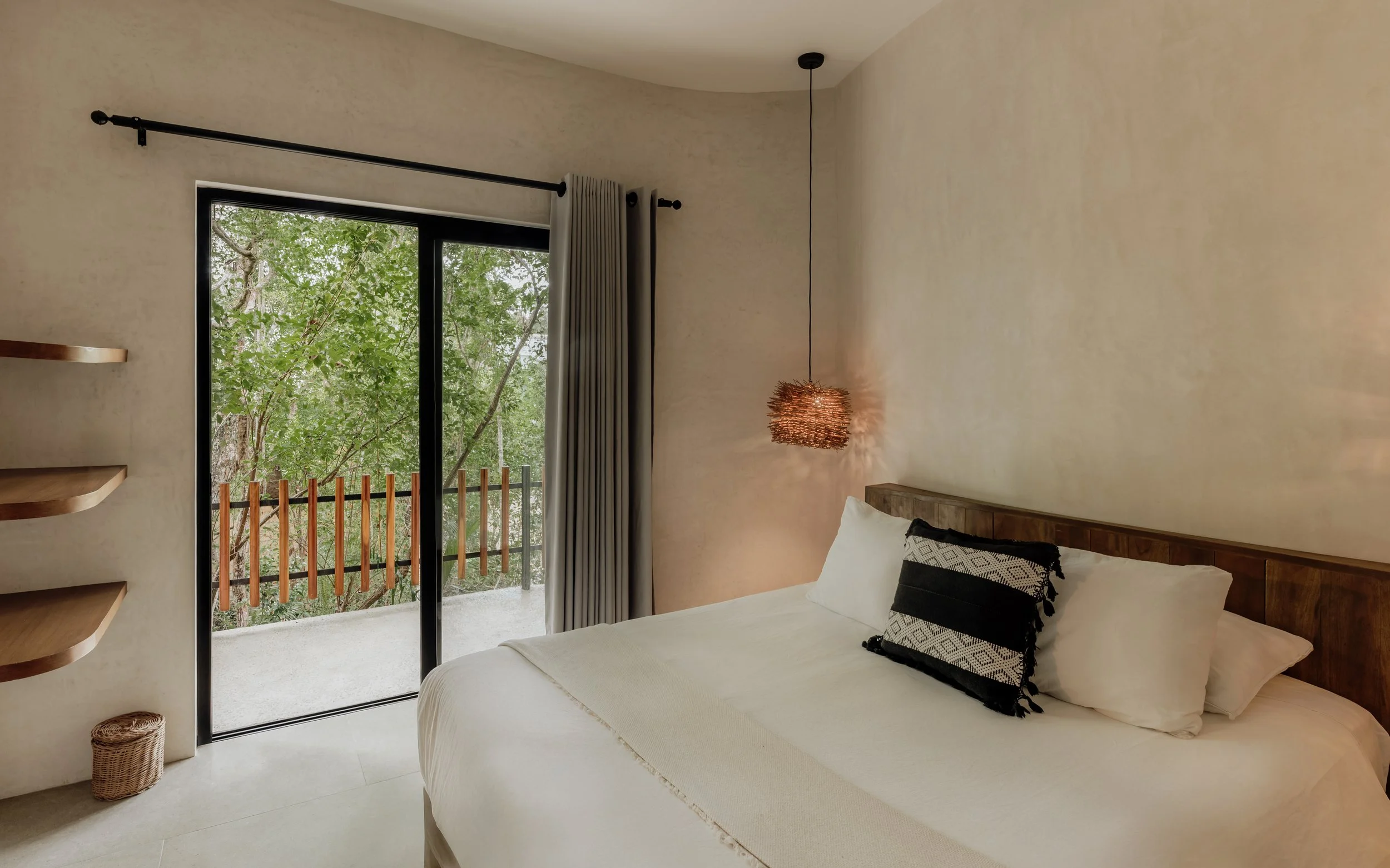 A bedroom with a bed, decorative pillows, wooden headboard, and a large window with curtains. Outside, trees are visible. There's a hanging pendant light and built-in shelves on the left wall.