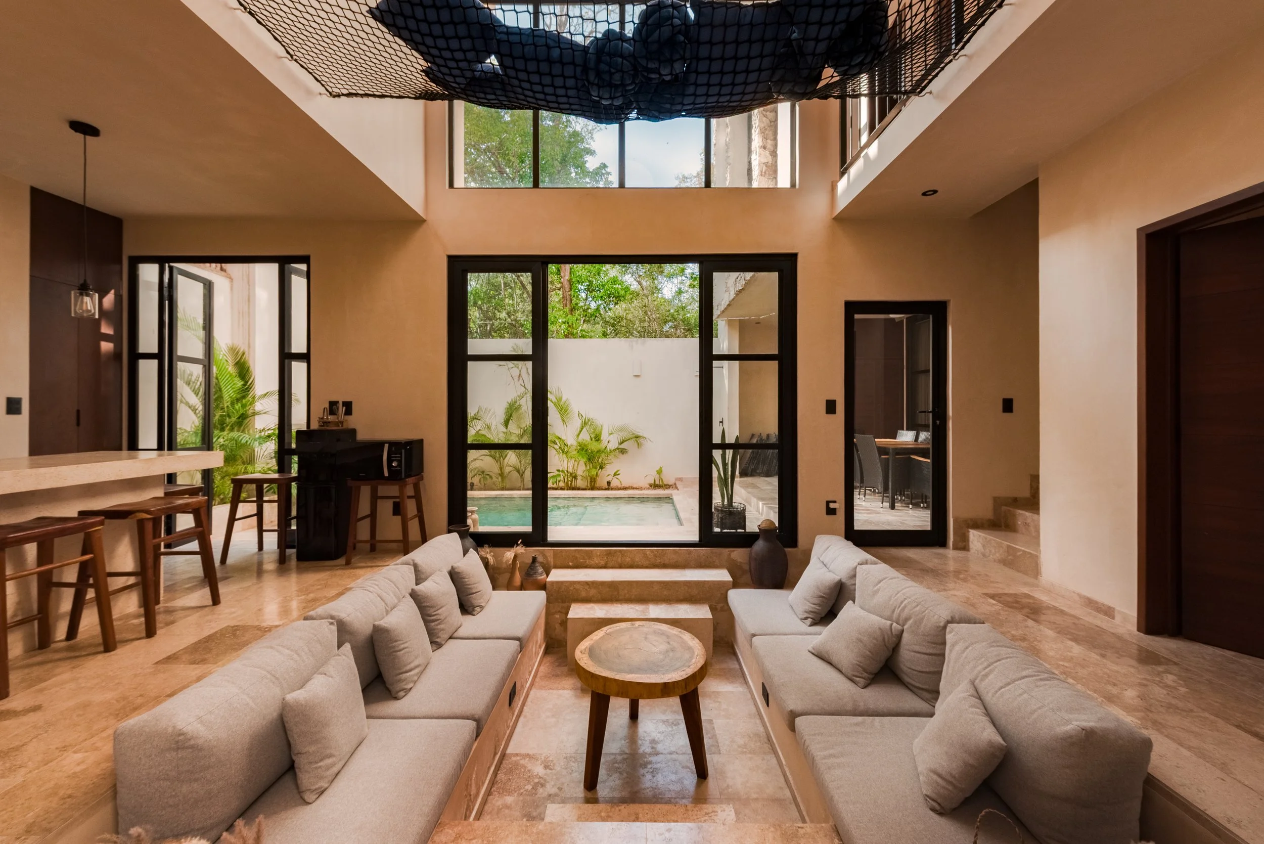 Living room with cream-colored sofas, wooden coffee table, large windows, and a view of outdoor greenery and pool.