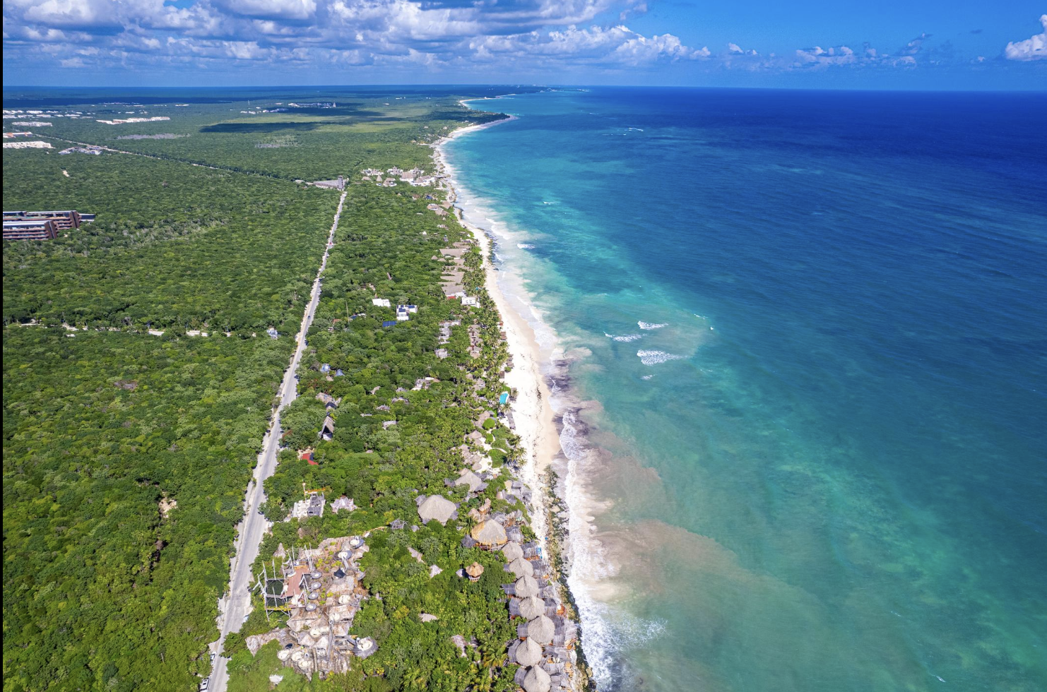 Aerial view of a tropical beach with turquoise water, white sandy shoreline, surrounded by lush green vegetation and scattered buildings.