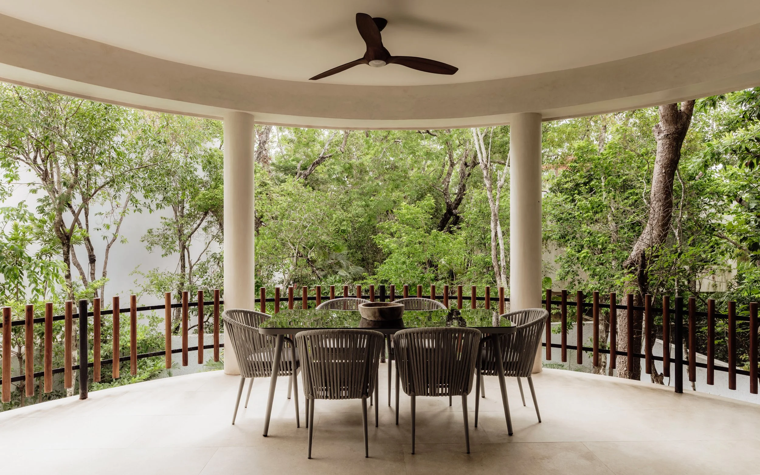 A covered outdoor patio with a round dining table, six chairs, and a ceiling fan, overlooking lush green trees and a white wall with a wooden railing.