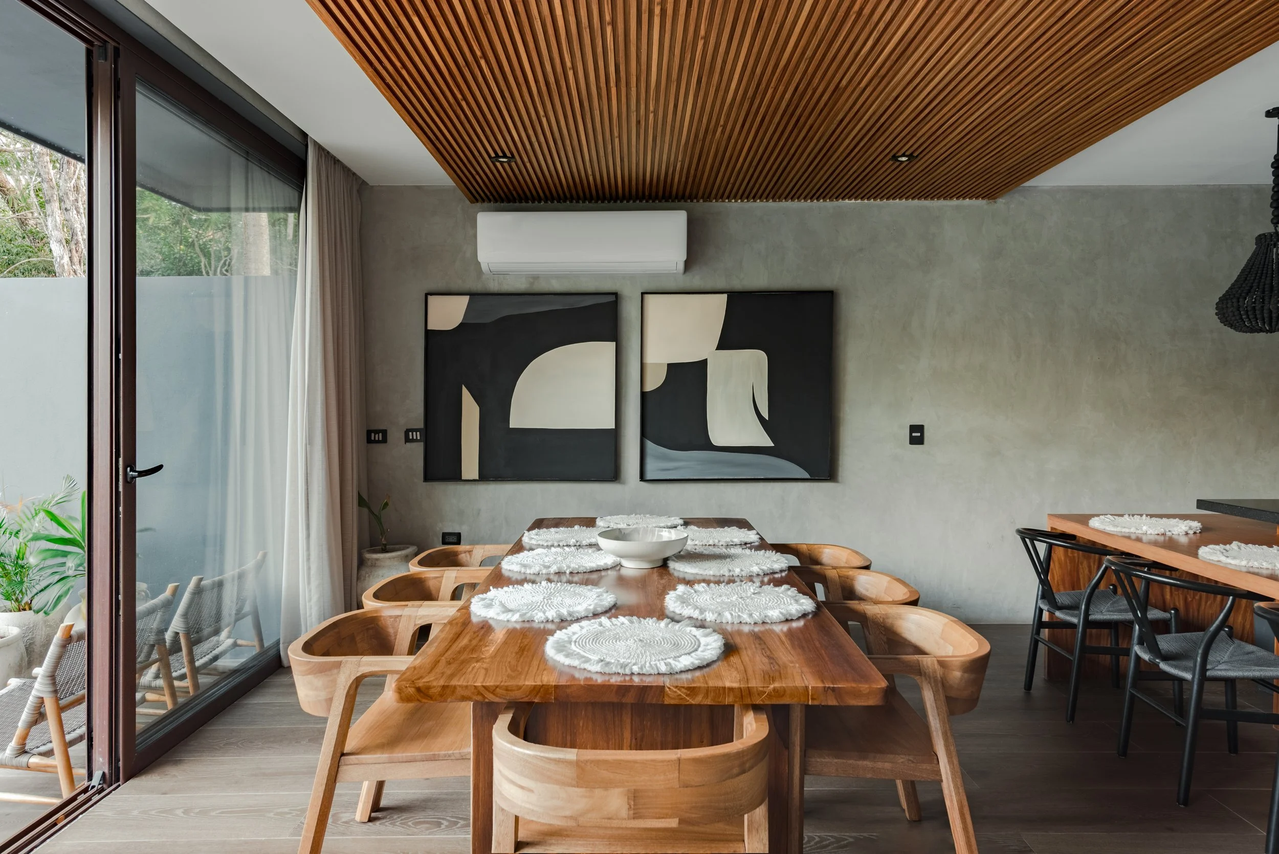A modern dining room with a large wooden table set with white woven placemats and a bowl, surrounded by matching wooden chairs. The room features a slatted wooden ceiling, artwork on the wall, large sliding glass doors with curtains, and outdoor greenery visible outside.
