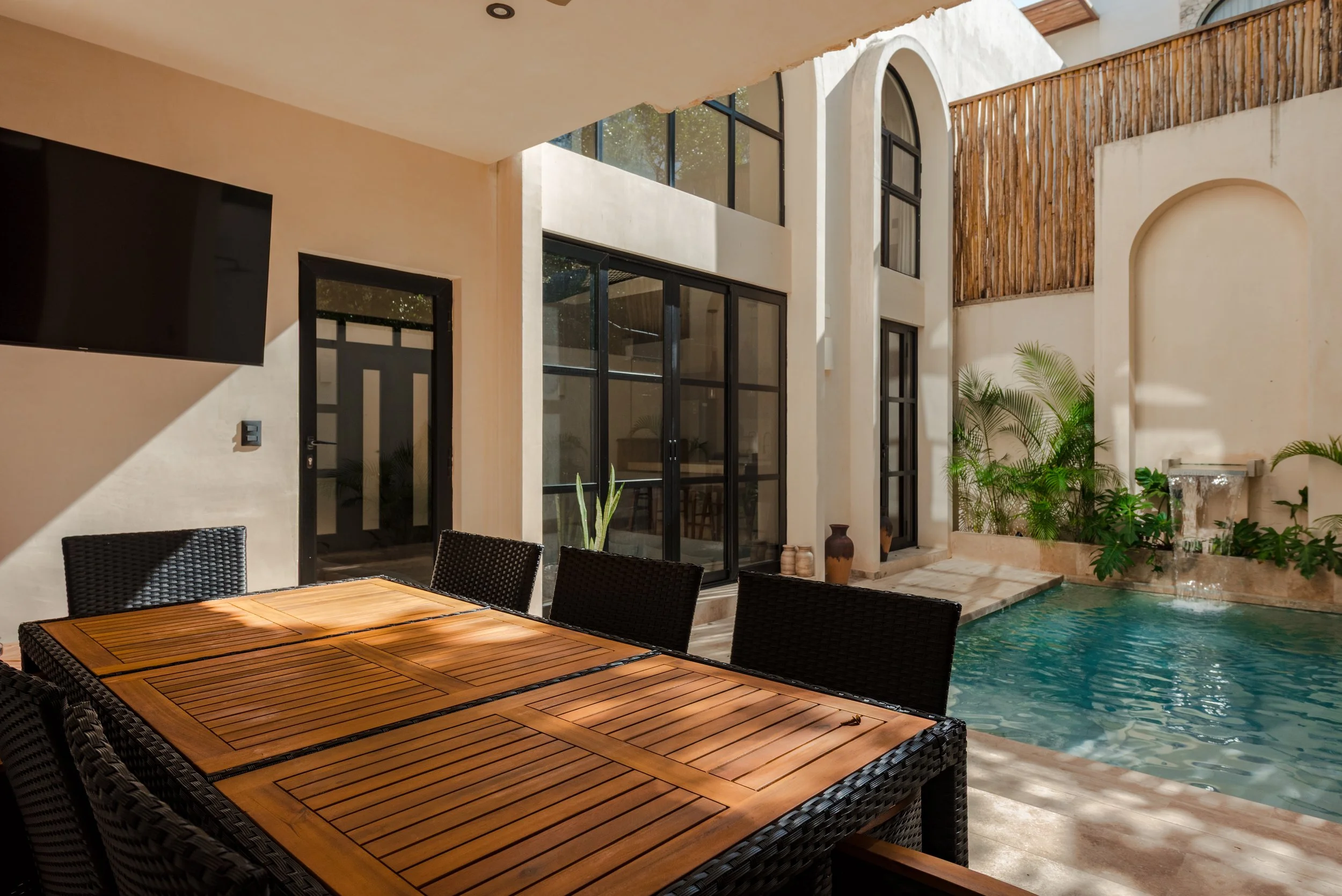 A modern indoor-outdoor living space with a wooden dining table, black wicker chairs, glass sliding doors, and a small pool surrounded by beige walls and green plants.