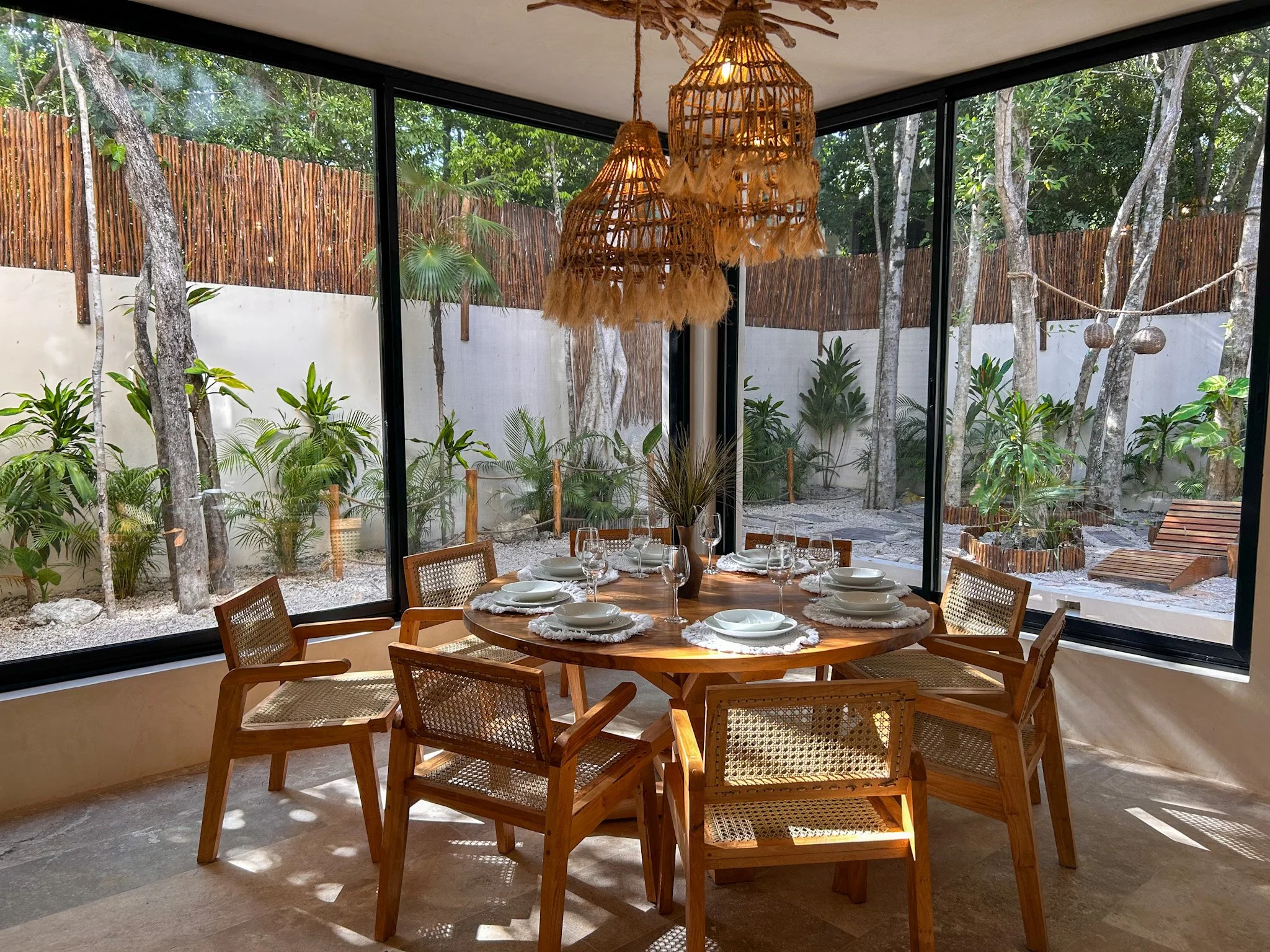 A dining area with a round wooden table set with white plates, glasses, and napkins, surrounded by wooden chairs with woven backs. Large glass windows reveal a lush outdoor garden with trees and greenery. A chandelier made of woven rattan hangs from the ceiling, and outside there is a bamboo fence and decorative hanging baskets.