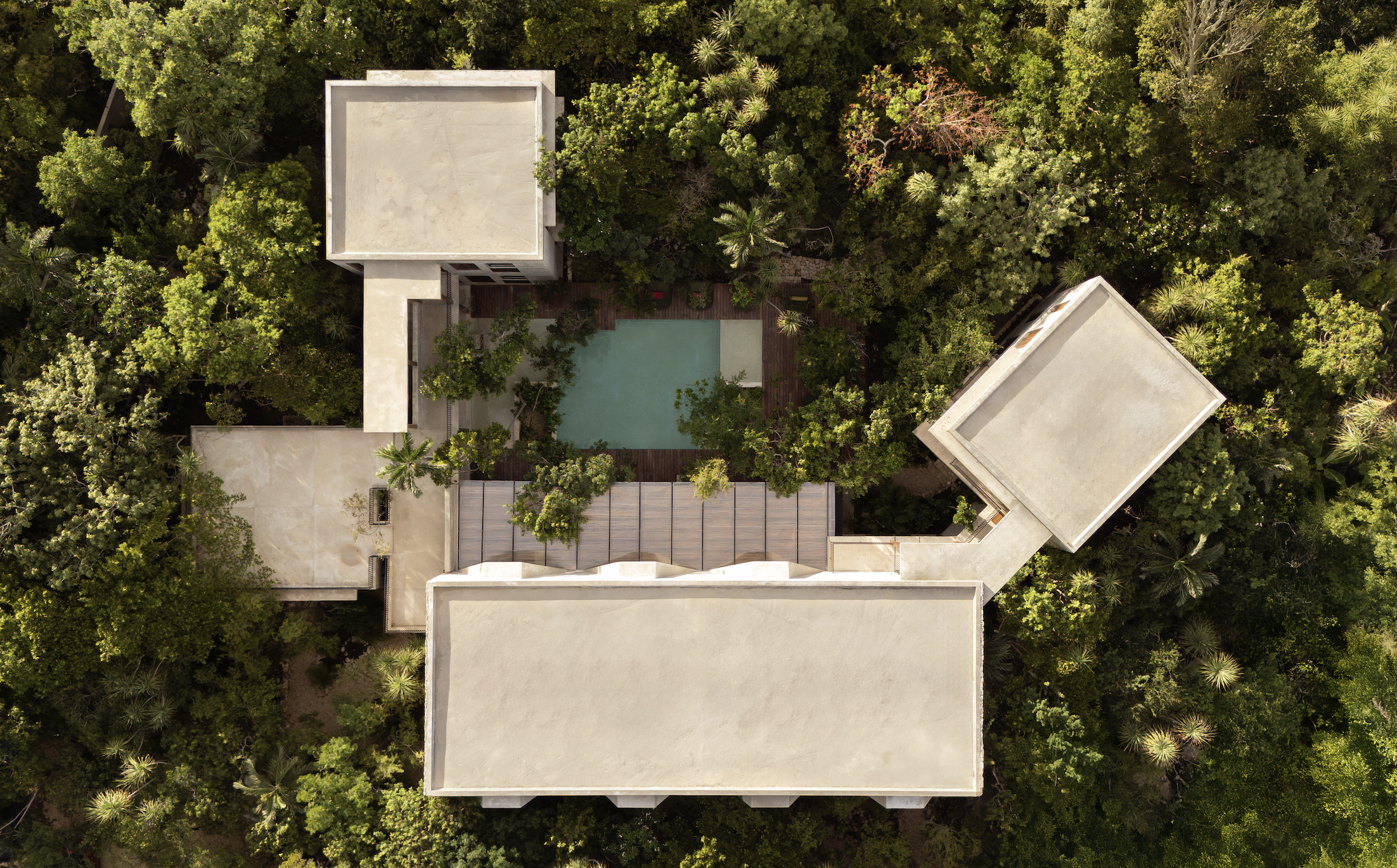 Aerial view of a modern building with a flat roof, surrounded by lush green trees and a small swimming pool with a wooden deck.