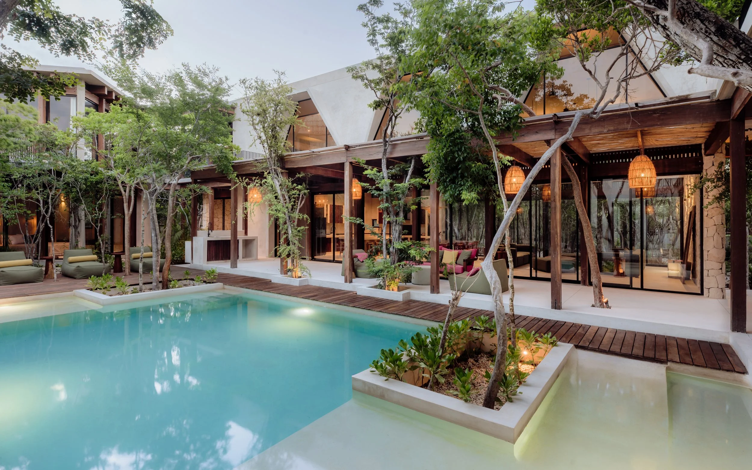 Modern house with large glass windows and a wooden deck, surrounded by trees and greenery, with a swimming pool in the foreground.