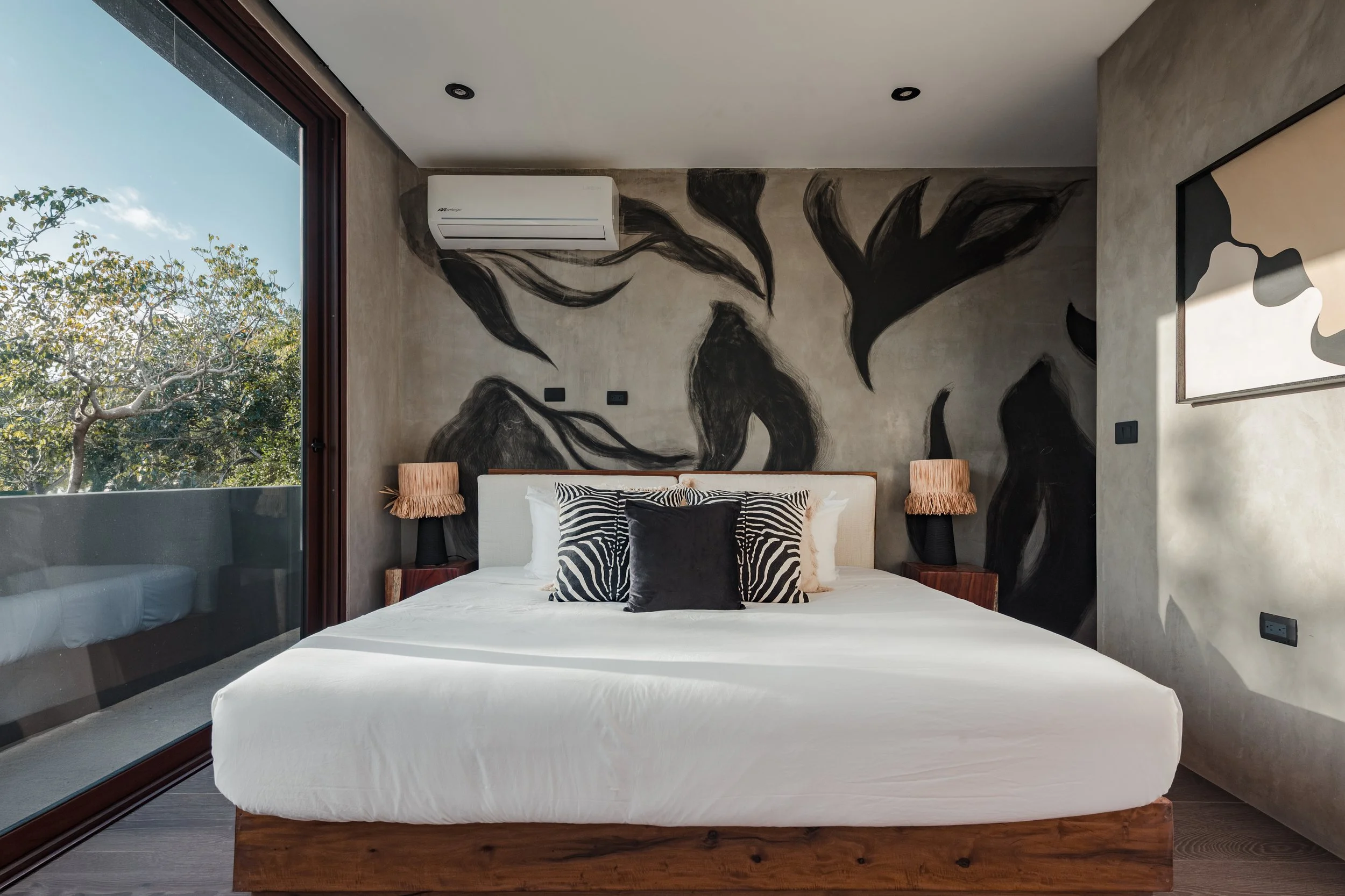 Modern bedroom with large window showing trees outside, featuring a sculptural black and white mural, a white bed with zebra pattern pillows, and minimalist decor.