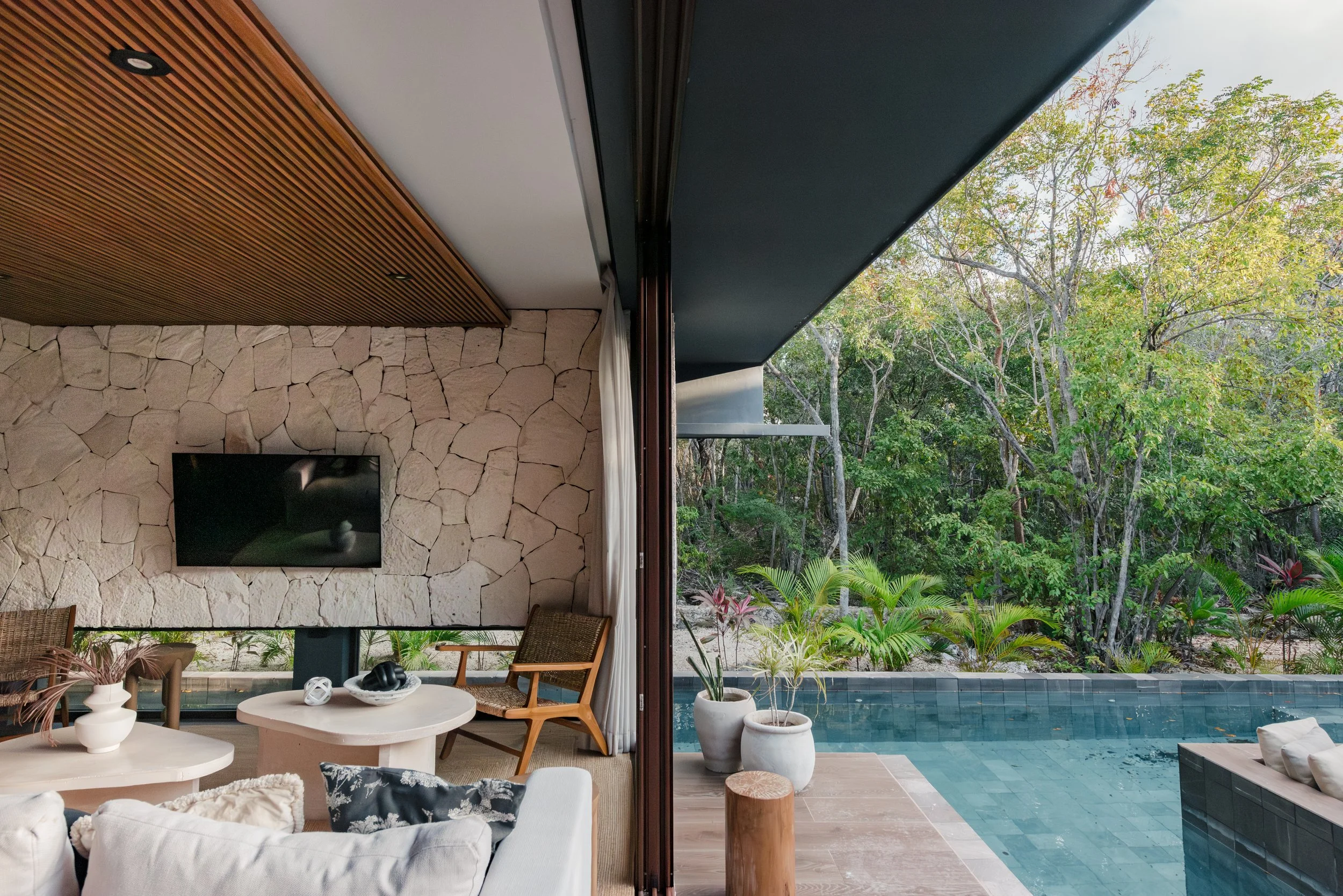 Interior of a modern house with a stone wall and a TV, opening to an outdoor pool and lush green trees.
