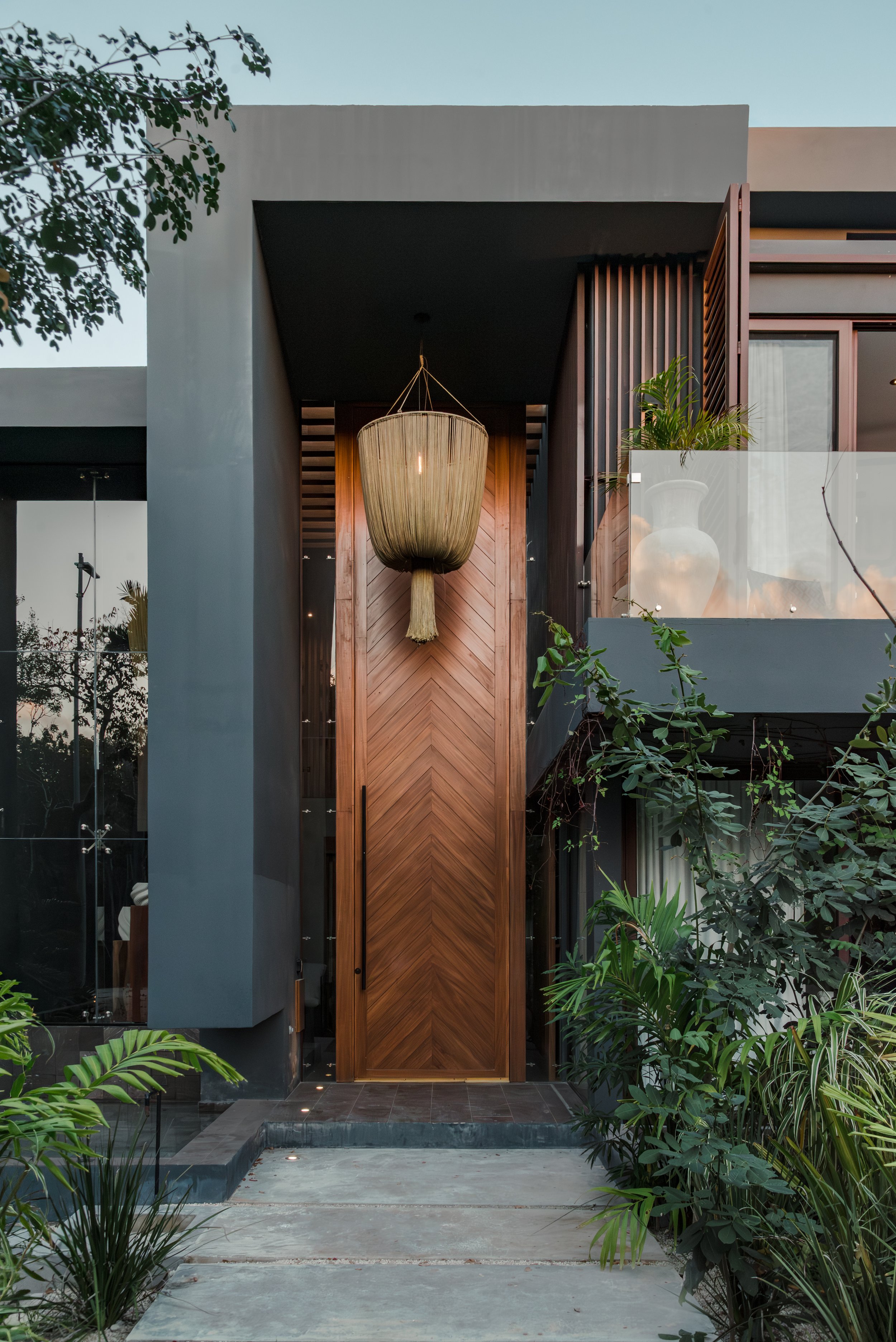 Modern house entrance featuring a large wooden door with chevron pattern, hanging woven light fixture, lush green plants, and a small pond with stepping stones.