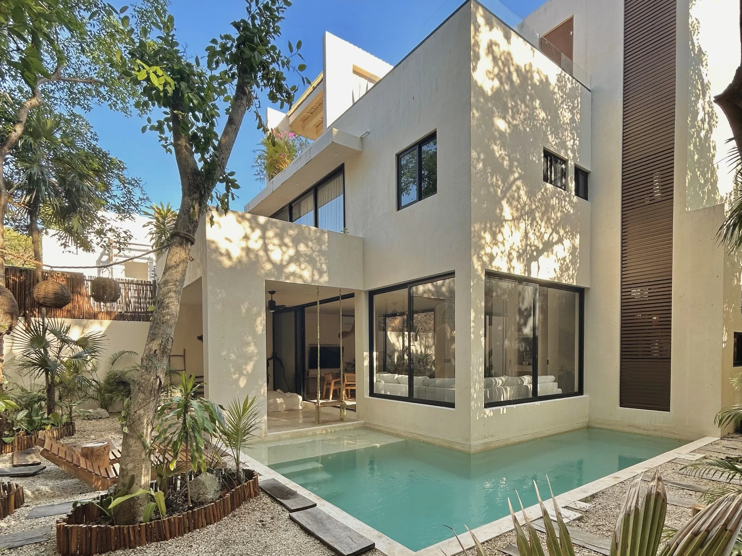 Modern white house with large floor-to-ceiling windows, surrounded by trees and greenery, with a swimming pool in the backyard.