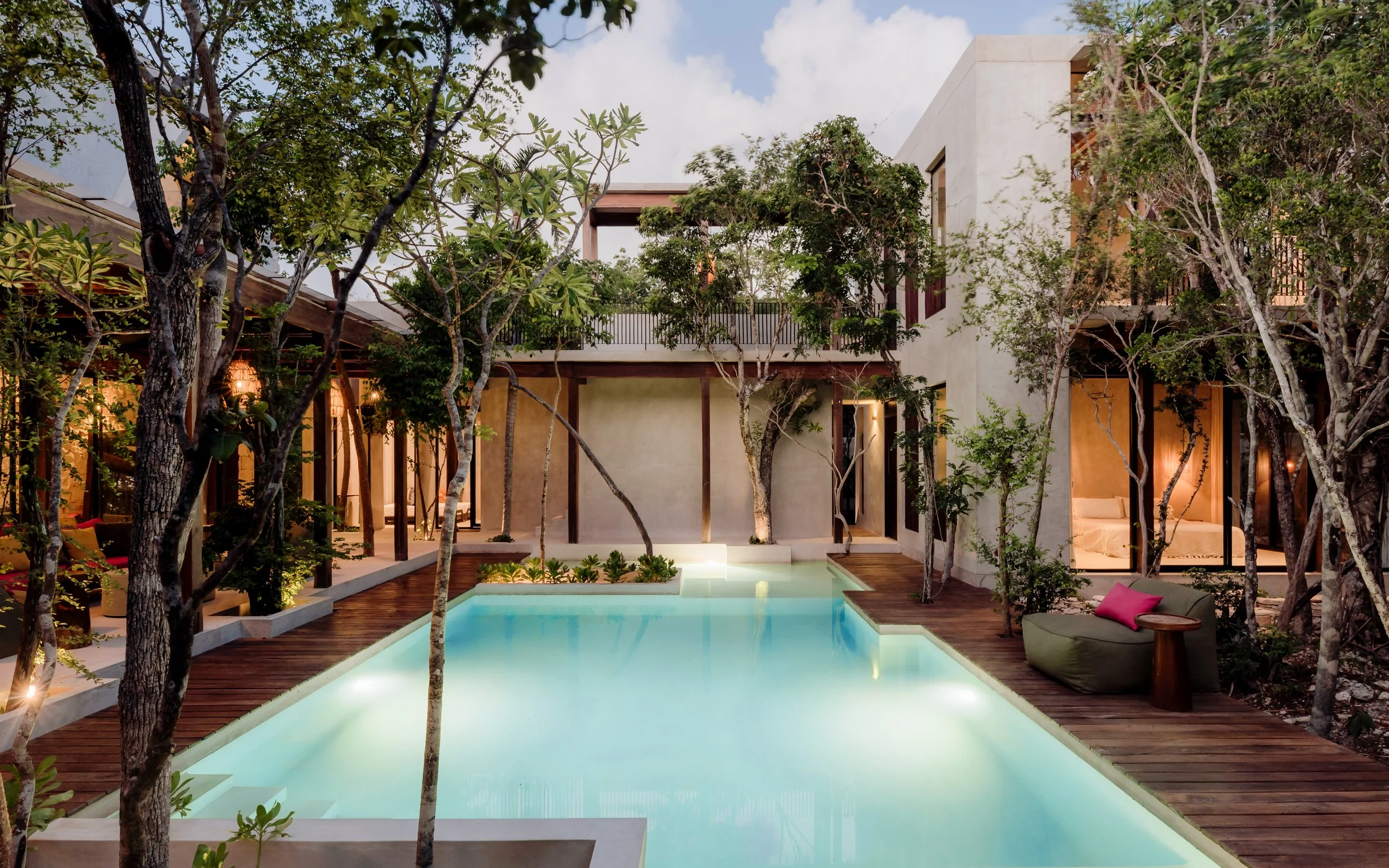 A modern house with a rectangular pool in the backyard, surrounded by trees and wooden deck. The house has large windows and outdoor seating with cushions, and is lit warmly from inside, creating a cozy atmosphere.