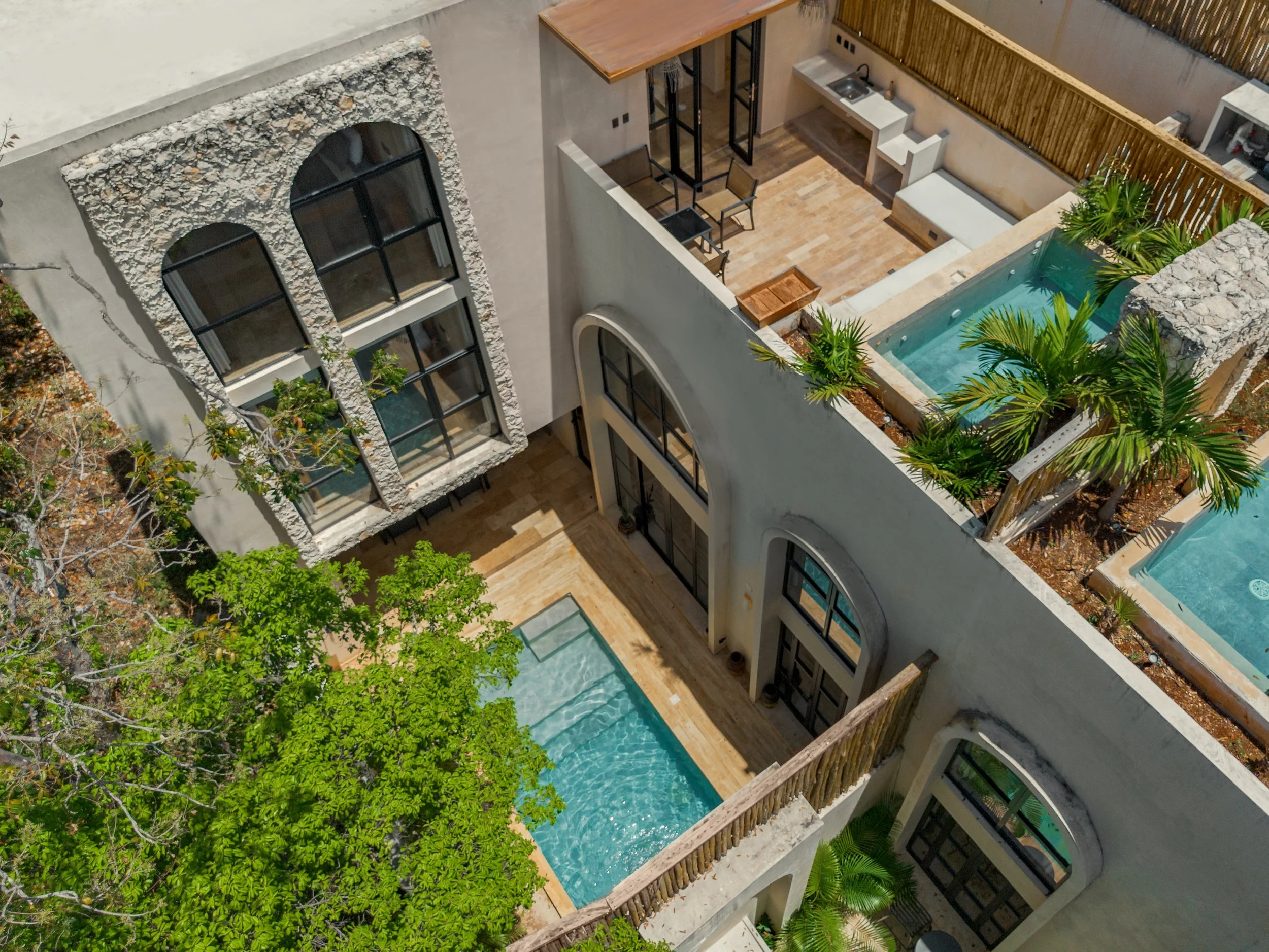 Aerial view of a modern residential building with large arched windows, a rooftop pool surrounded by wooden fencing, and lush green trees.