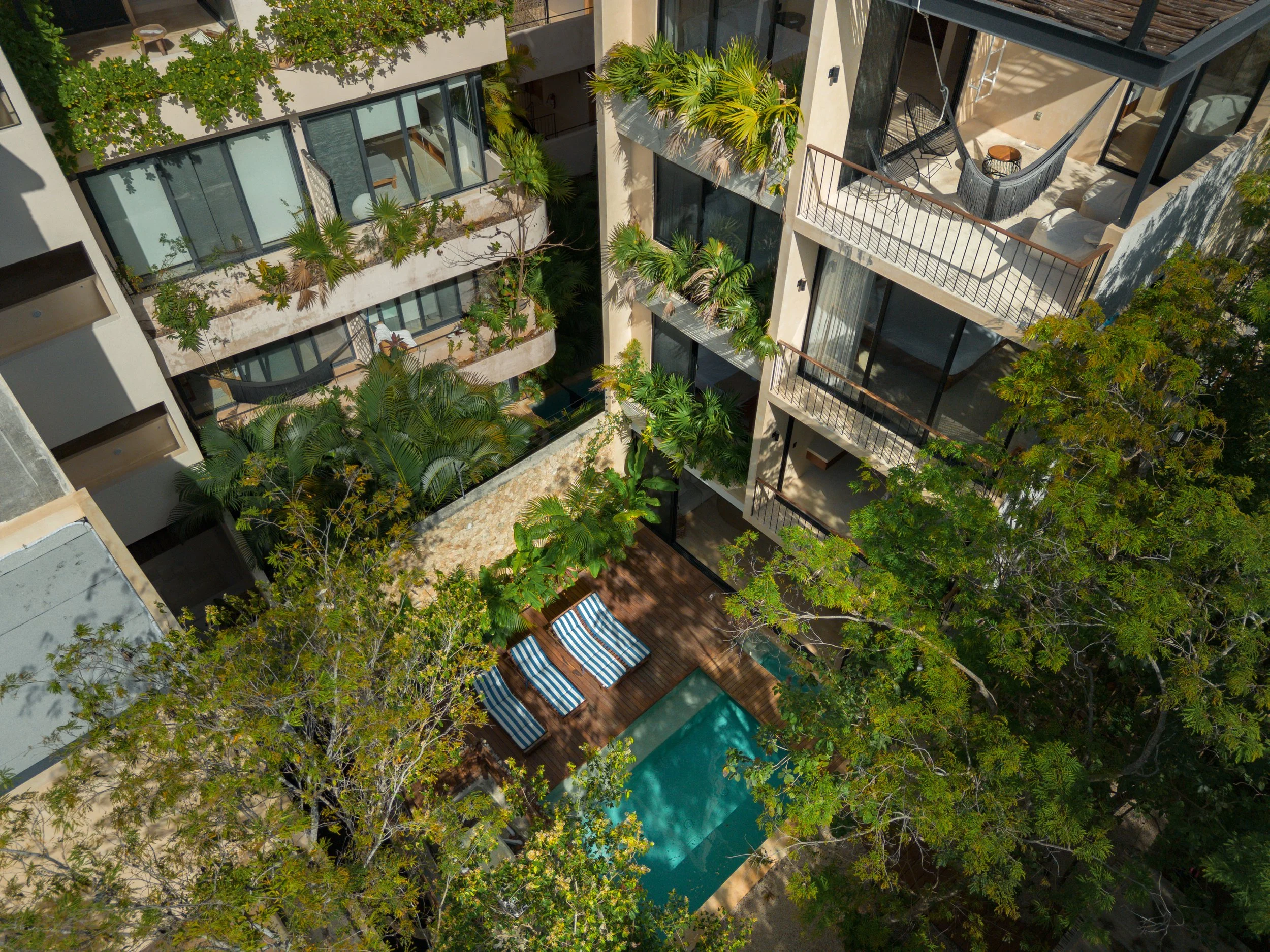 An aerial view of a multi-story modern apartment building with balconies, lush green trees, a swimming pool, and outdoor lounge furniture.