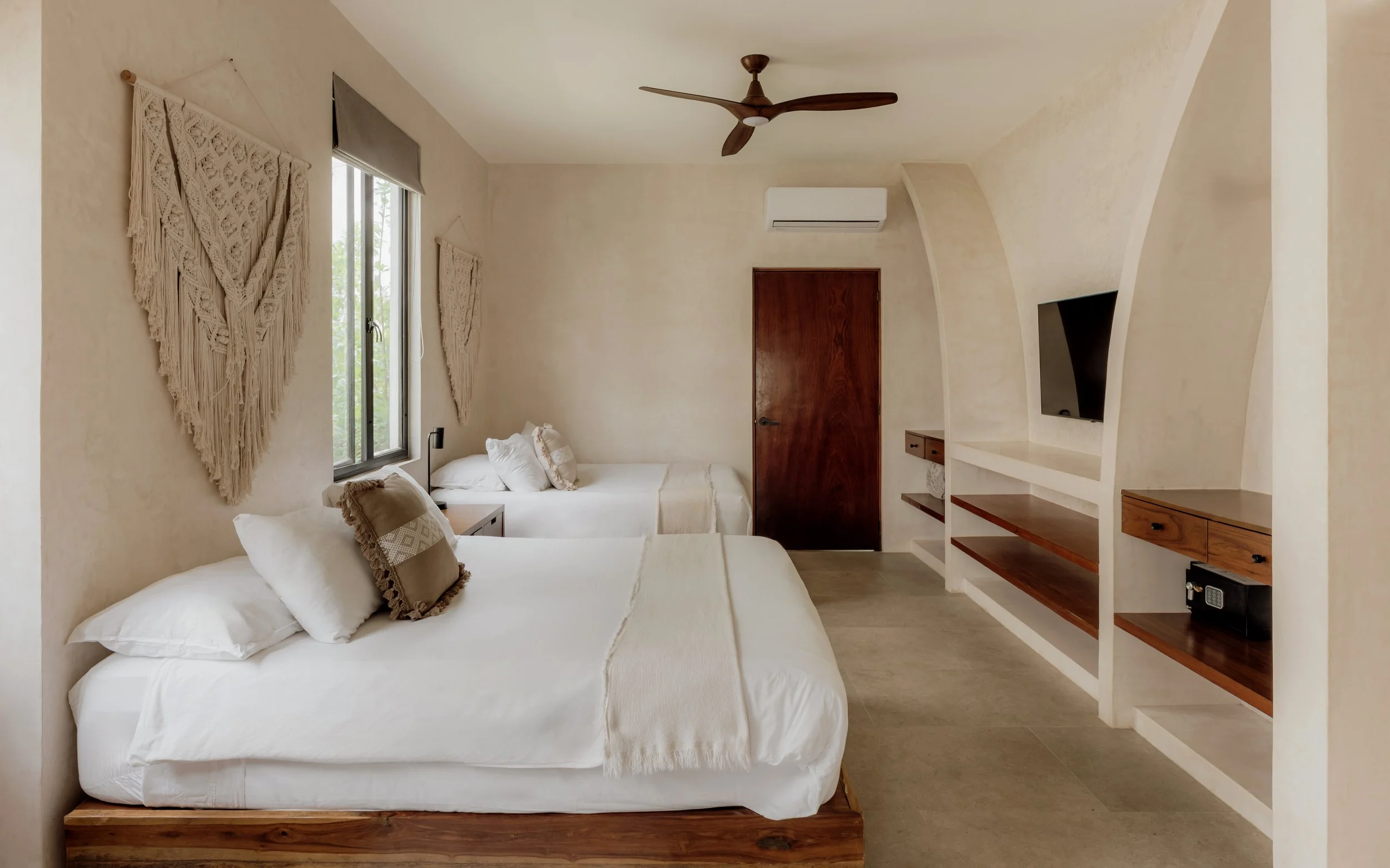 A room with two neatly made beds, decorative wall hangings, a window with blinds, a wooden door, a flat-screen TV embedded in built-in shelves, and a ceiling fan.