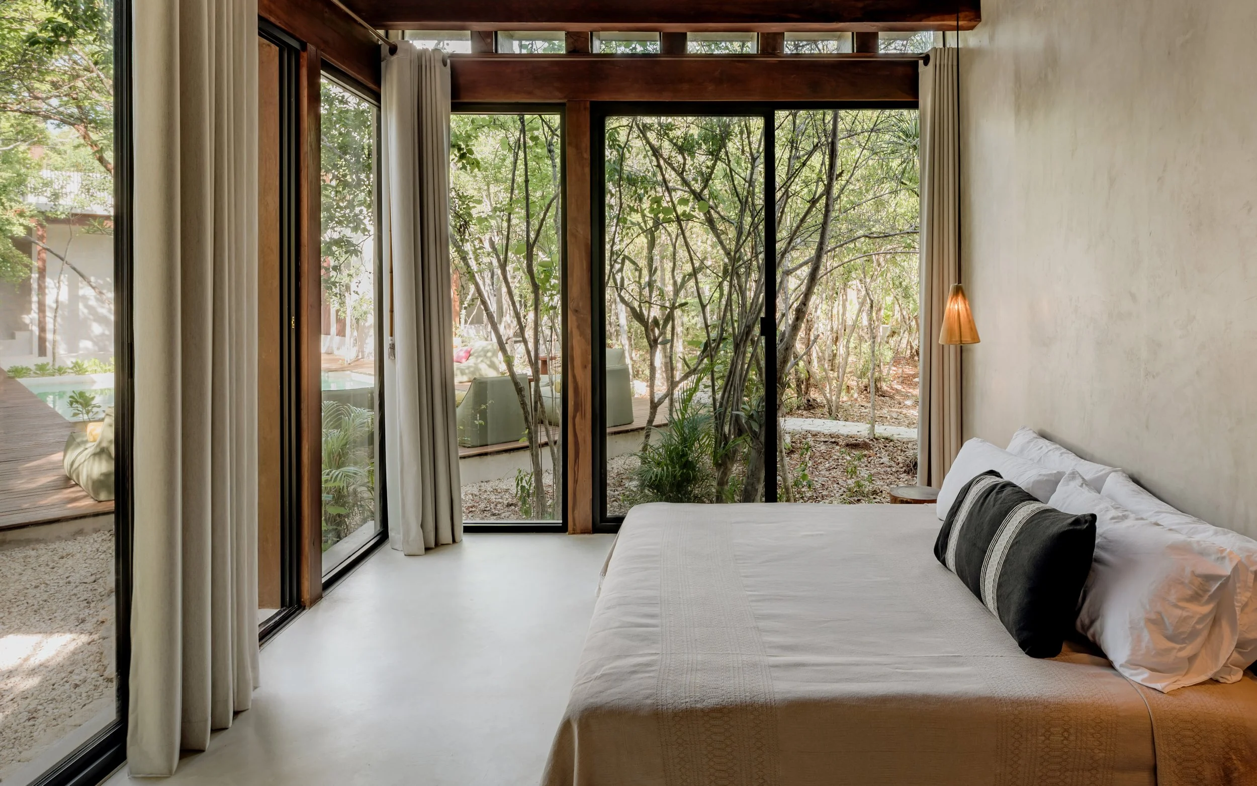 Interior view of a bedroom with a large glass sliding door showing trees outside, a bed with white linens and pillows, and a small orange bedside lamp.