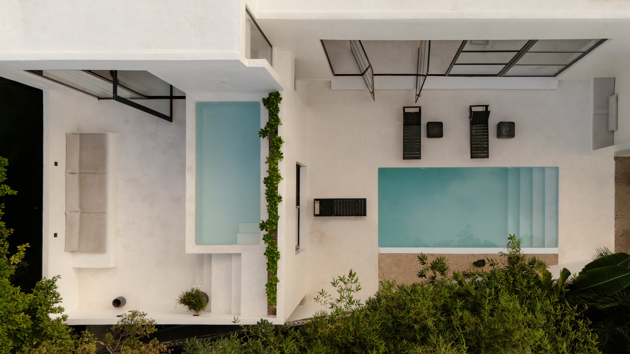 Aerial view of a modern backyard with two swimming pools, black lounge chairs, white walls, and surrounding greenery.
