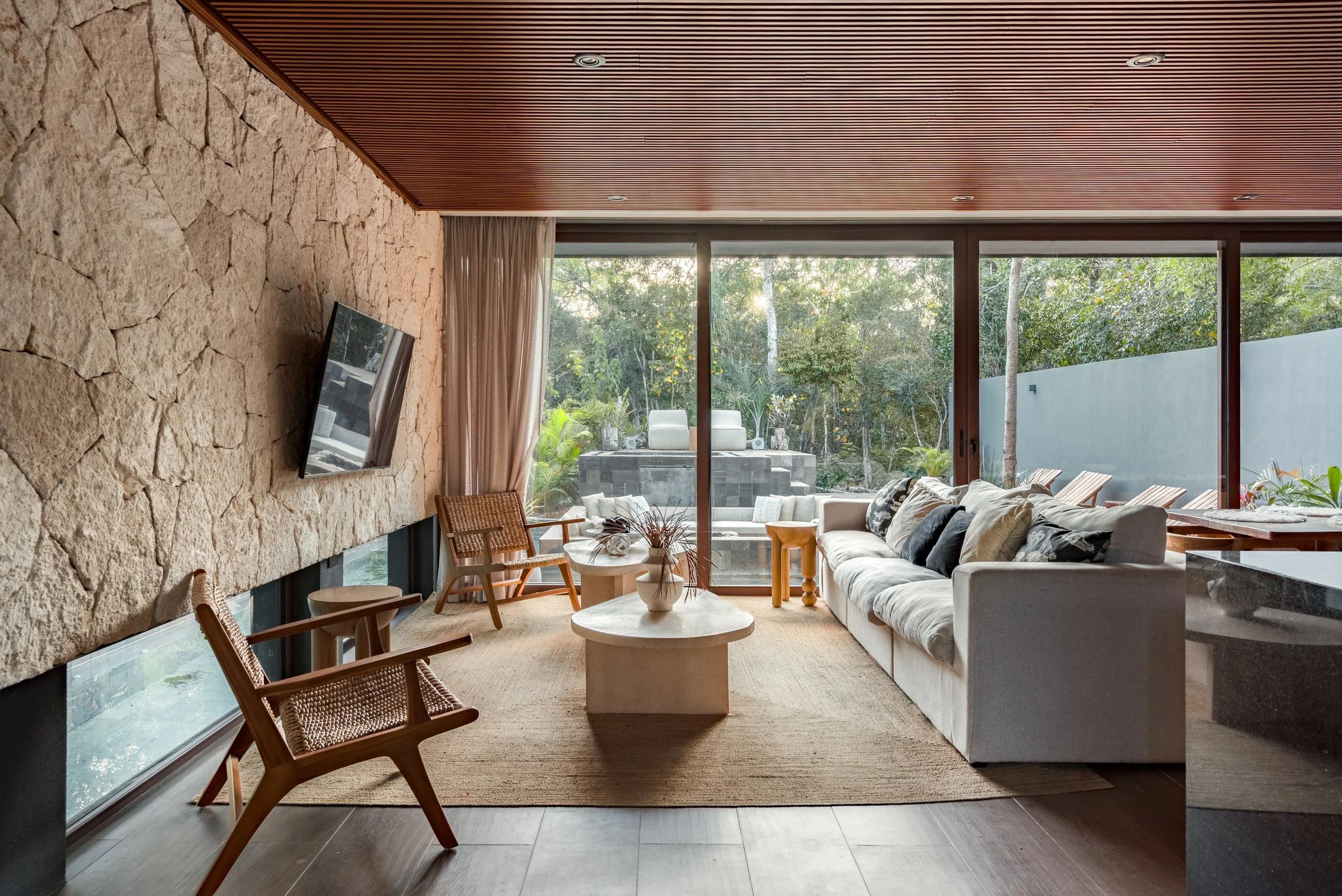 Modern living room with large floor-to-ceiling windows overlooking greenery, stone accent wall, white sofa with black and white pillows, round coffee table, wooden chairs, beige rug, and ceiling with wooden slats and recessed lighting.