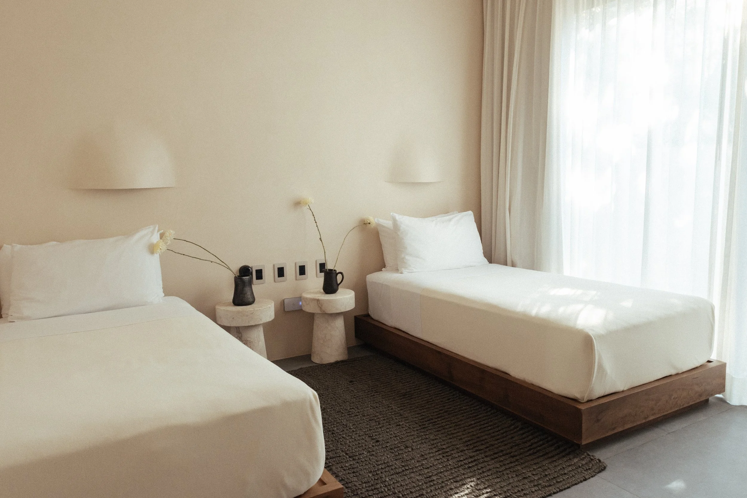A minimalist bedroom with two single beds, each with white bedding and pillows, a small round marble side table between them holding black vases, and light-colored curtains letting in natural light.