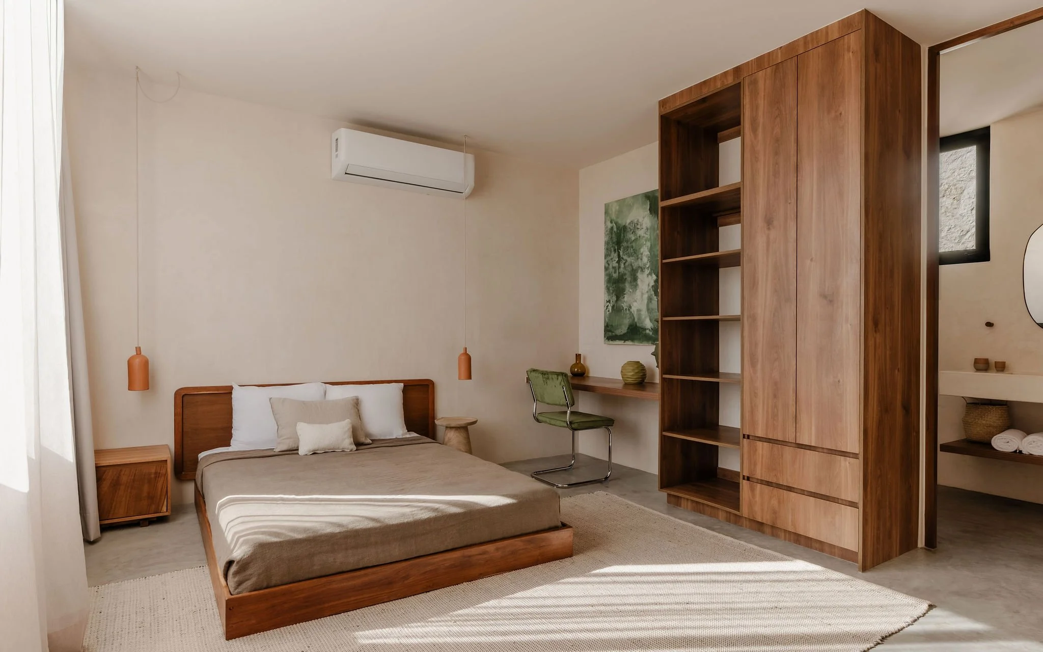 A bedroom with a wooden bed frame, beige bedding, and pillows, flanked by matching wooden nightstands, and a large wooden shelving unit dividing the space, with artwork, a green chair, and decorative items visible.