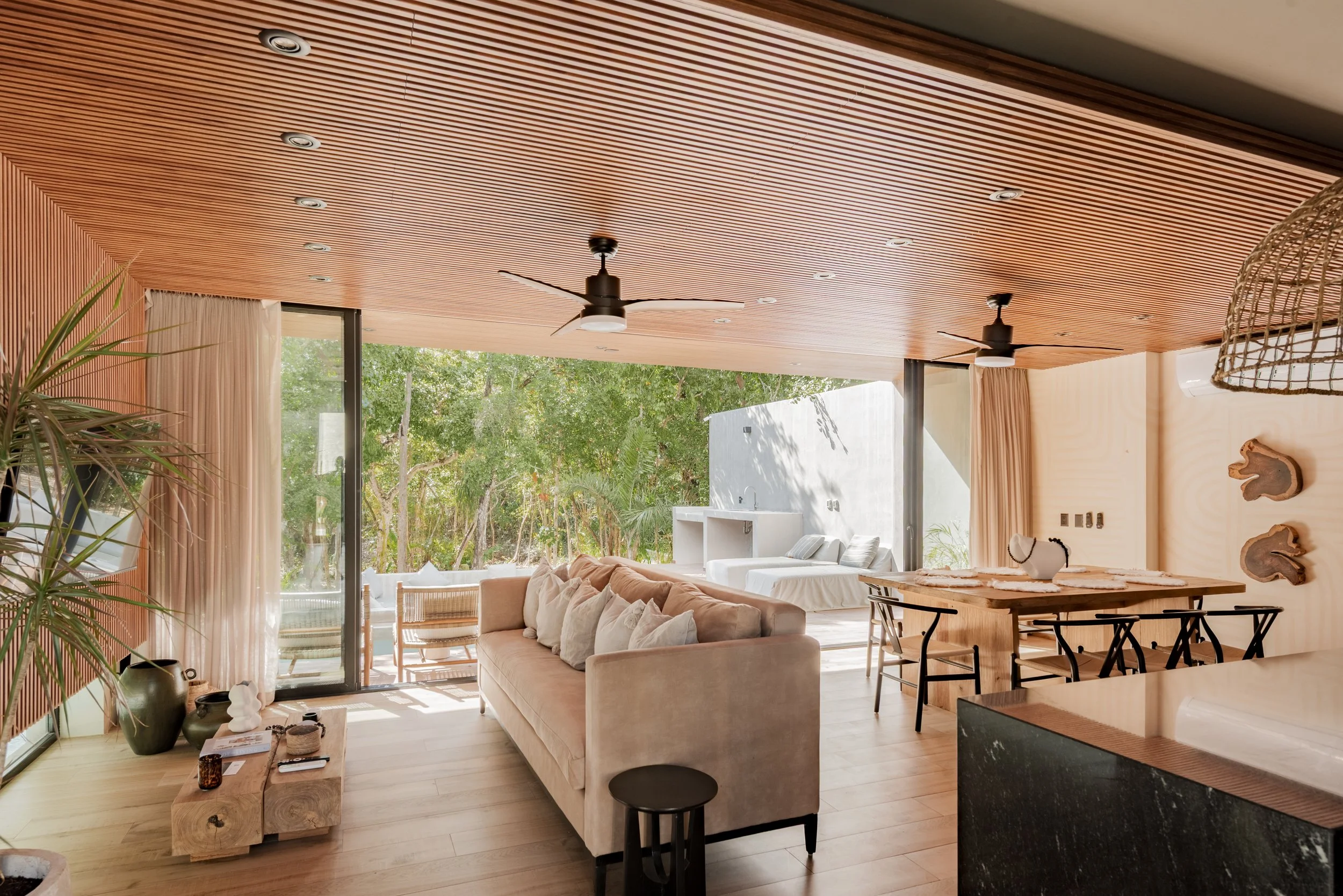 Interior of a modern living room with a wooden ceiling featuring embedded lights and ceiling fans, large glass windows with cream curtains, a beige sofa with white pillows, a wooden coffee table, indoor plants, and a view of trees outside.