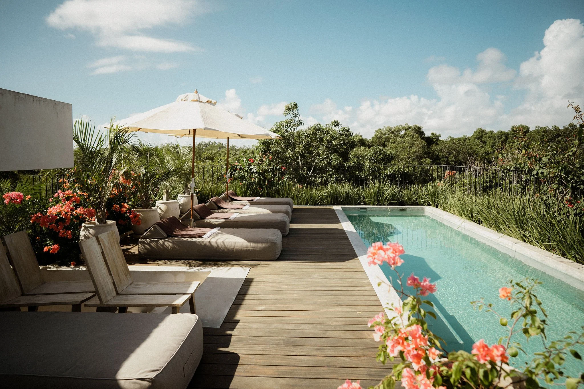 A backyard with a swimming pool, several lounge chairs with cushions and towels, an outdoor umbrella, lush green plants and flowers, and a clear blue sky with some clouds.