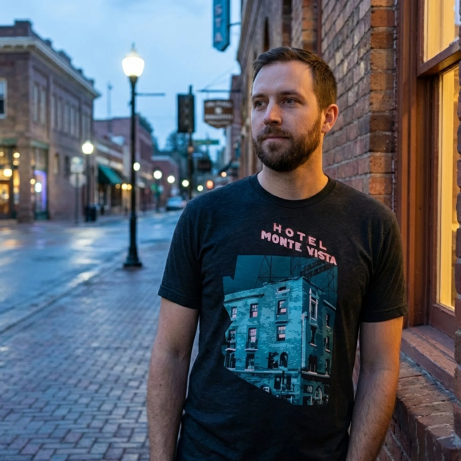 A man with short dark hair and a beard stands on a city street at dusk, wearing a black T-shirt with a graphic of a building and the words 'Hotel Monte Vista' on it. The street is wet and lined with brick buildings with lit windows, street lamps, and signs.