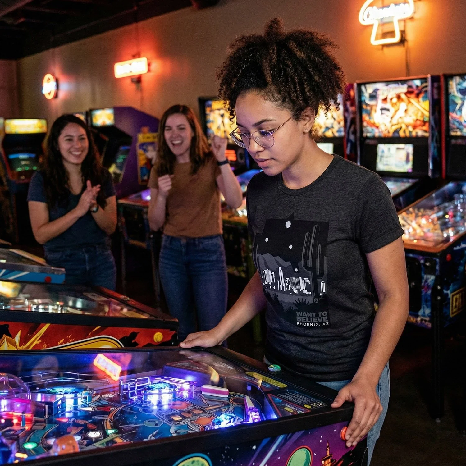 A young woman playing pinball at an arcade, with two girls cheering her on in the background.