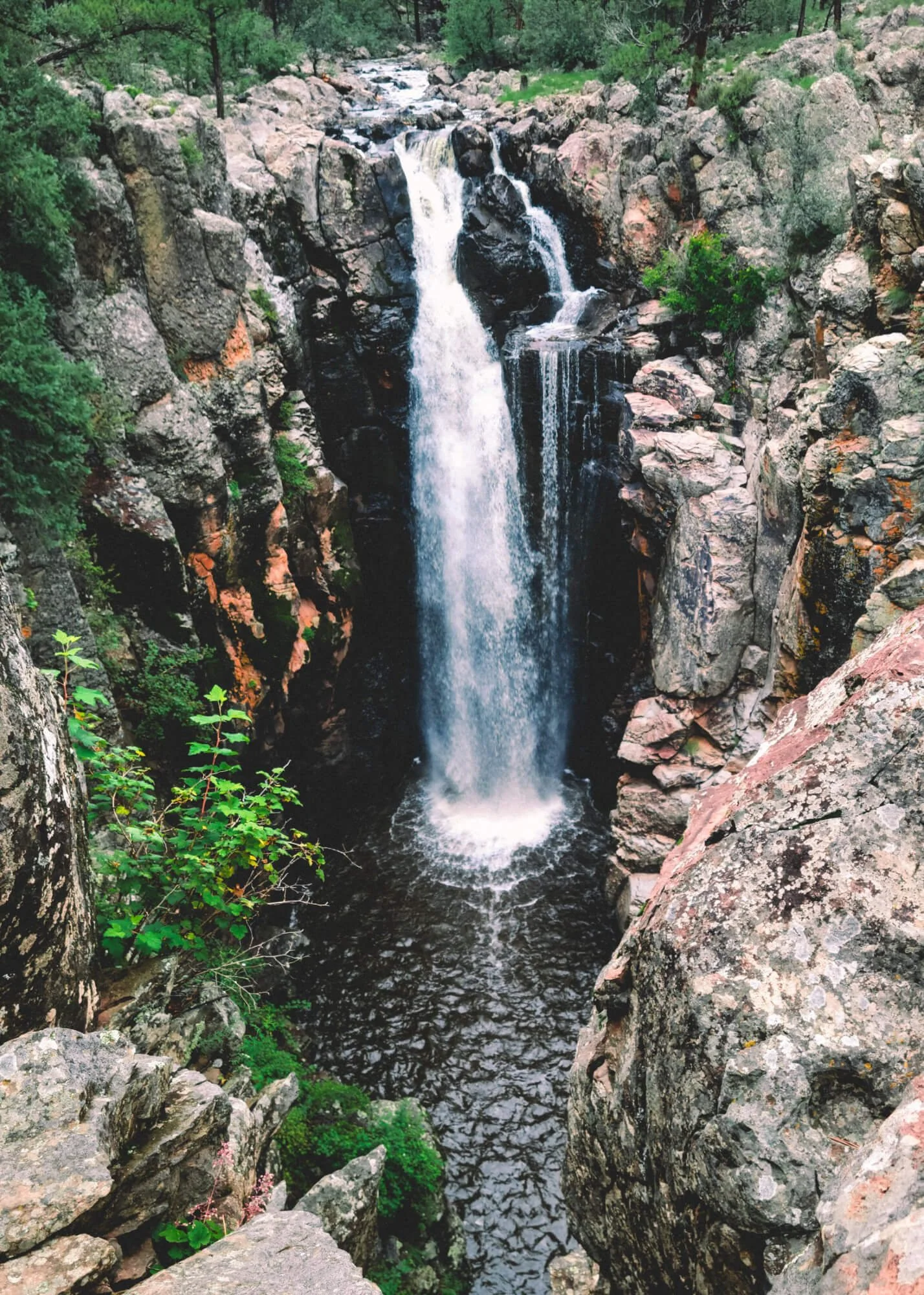 A tall waterfall cascading down rocks into a pool below, surrounded by green trees and rugged cliffs.