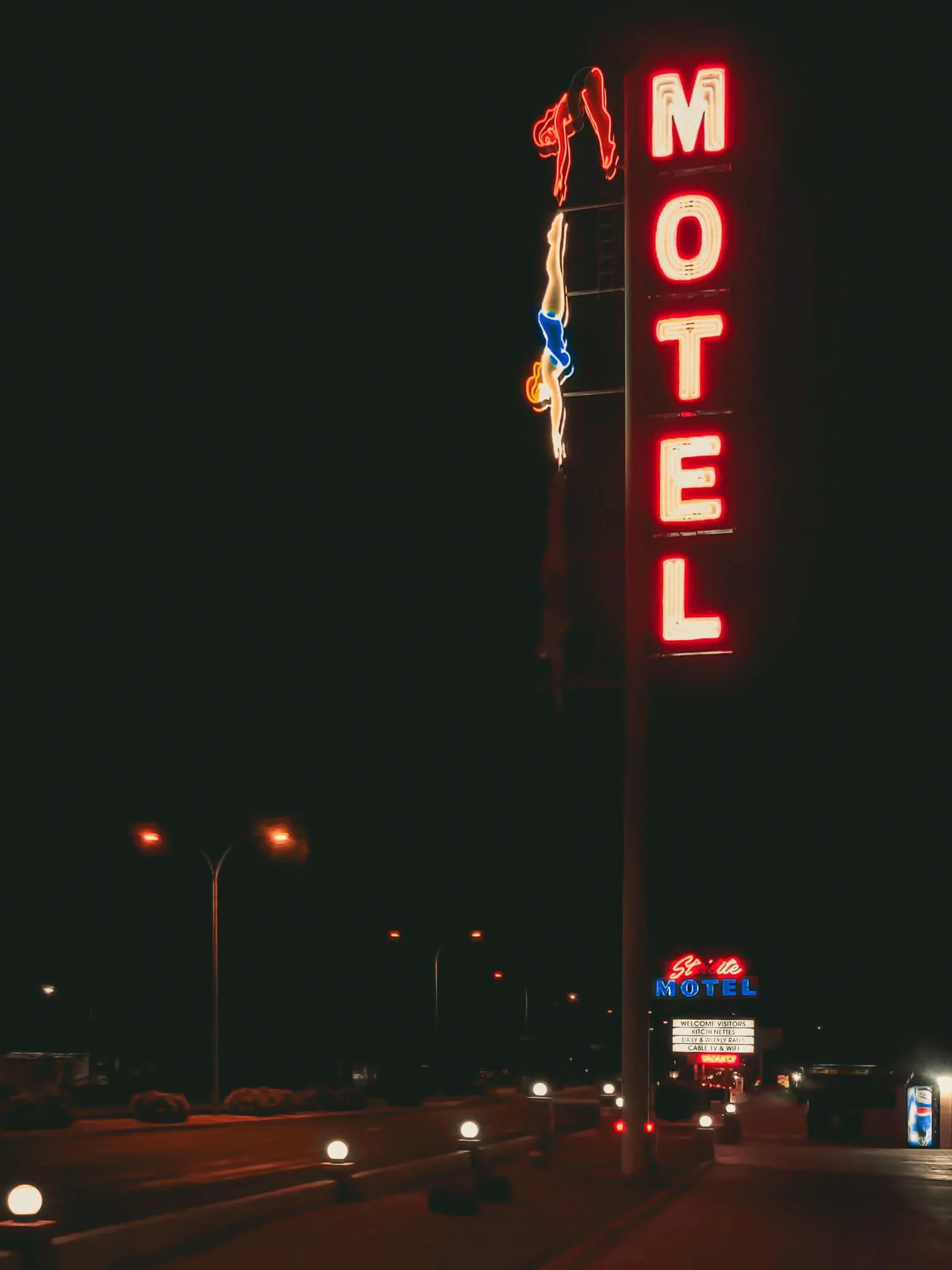 Night view of a neon sign for 'MOTEL' in red lights with a dancer figure on top, and a smaller sign below reading 'Sevilla MOTEL'.
