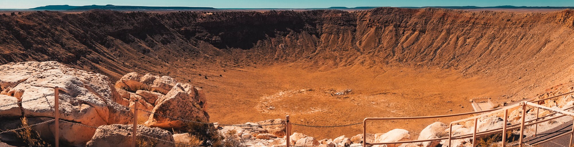 A large crater with rocky sides and a flat, barren center surrounded by a guardrail and steps, with a distant mountain range in the background.