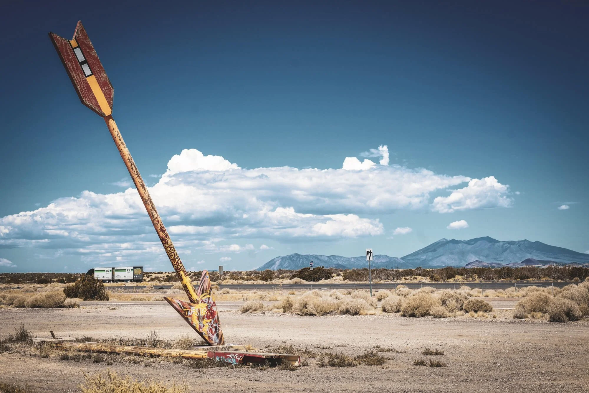 A toppled, graffiti-covered sign in a desert landscape with mountains and clouds in the background.