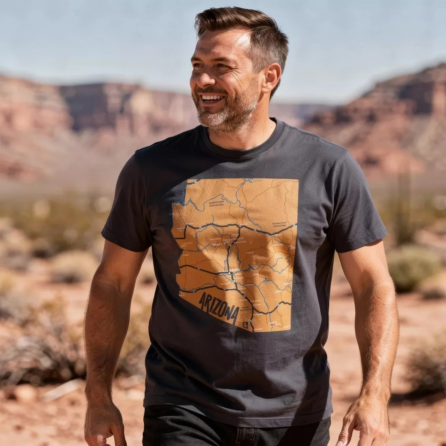 A man smiling outdoors in a desert landscape, wearing a black T-shirt with a map of Arizona on it.