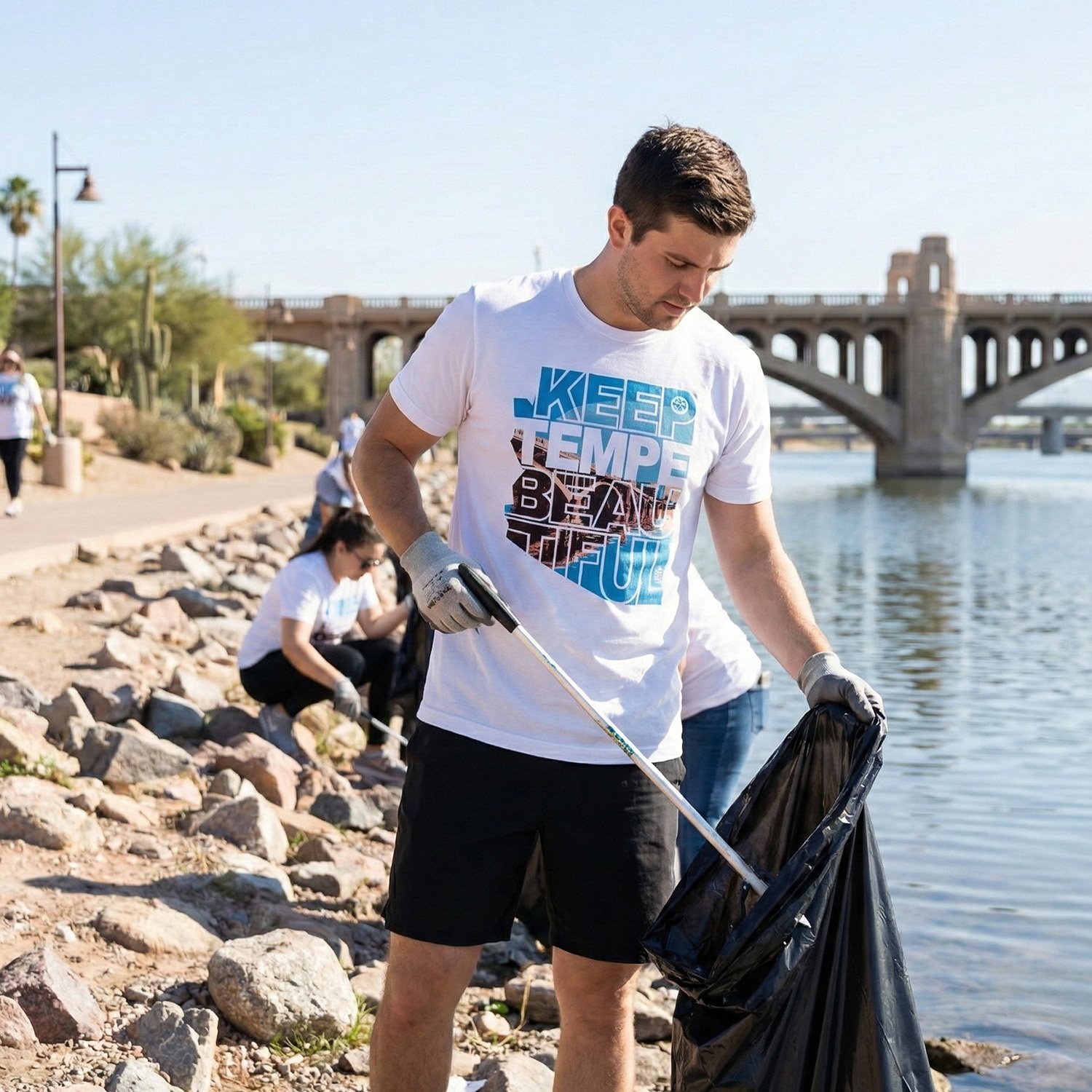 A young man in a white T-shirt and black shorts collecting trash along the water's edge with a group of people participating in a cleanup event.