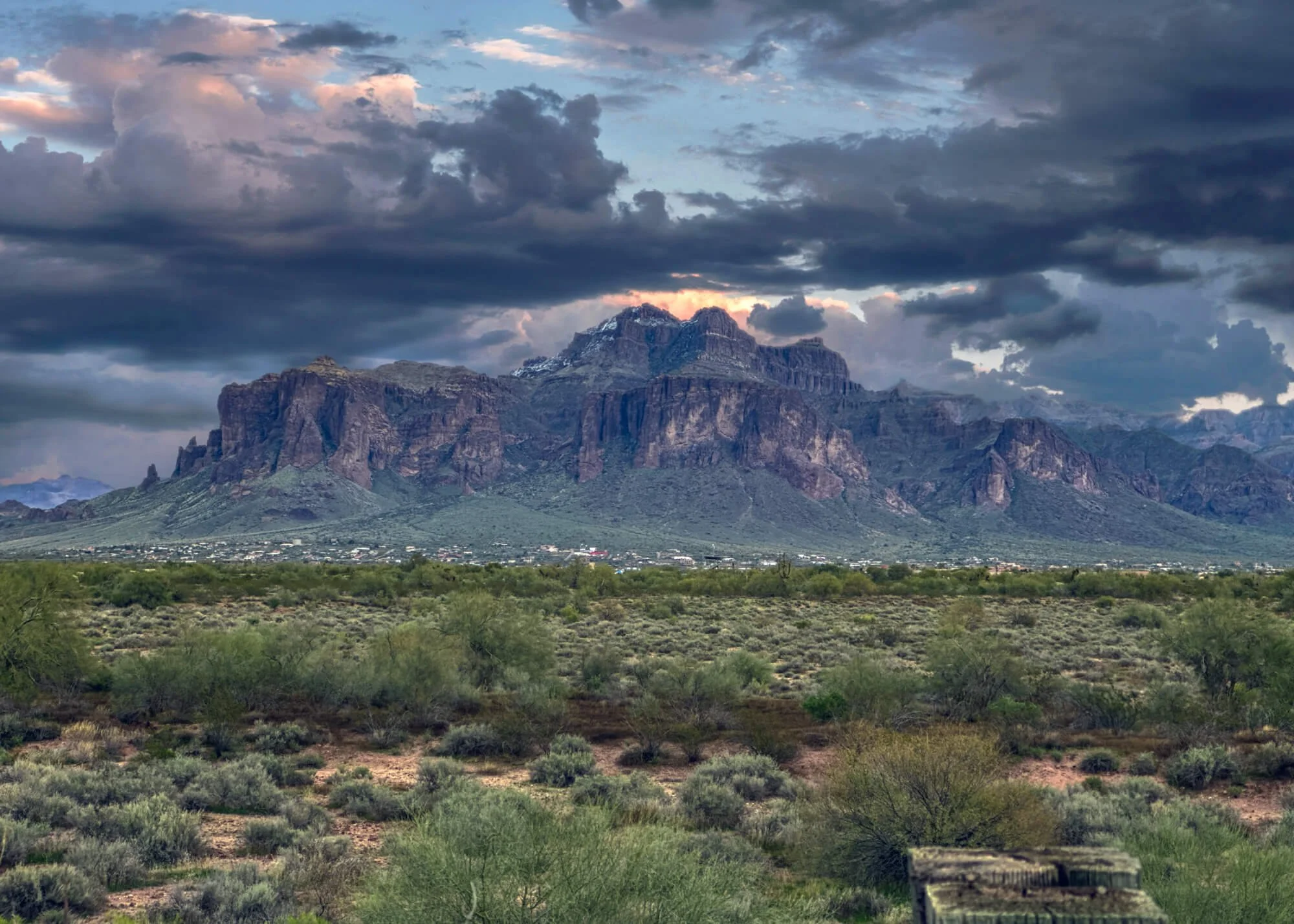 A large mountain range under a cloudy sky with some snow on peaks, and desert bushland in the foreground.