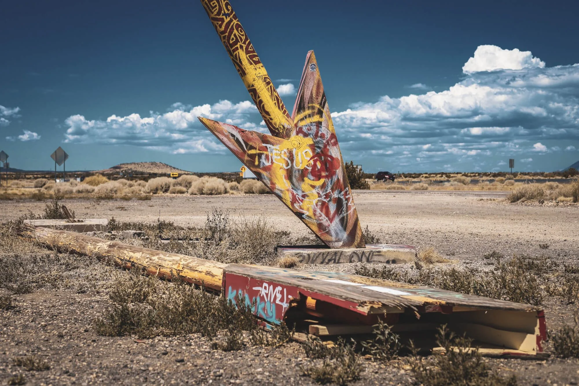 Graffiti-covered metal star sculpture tilted on the ground in a barren desert landscape with scattered bushes and a distant horizon under a blue sky with clouds.