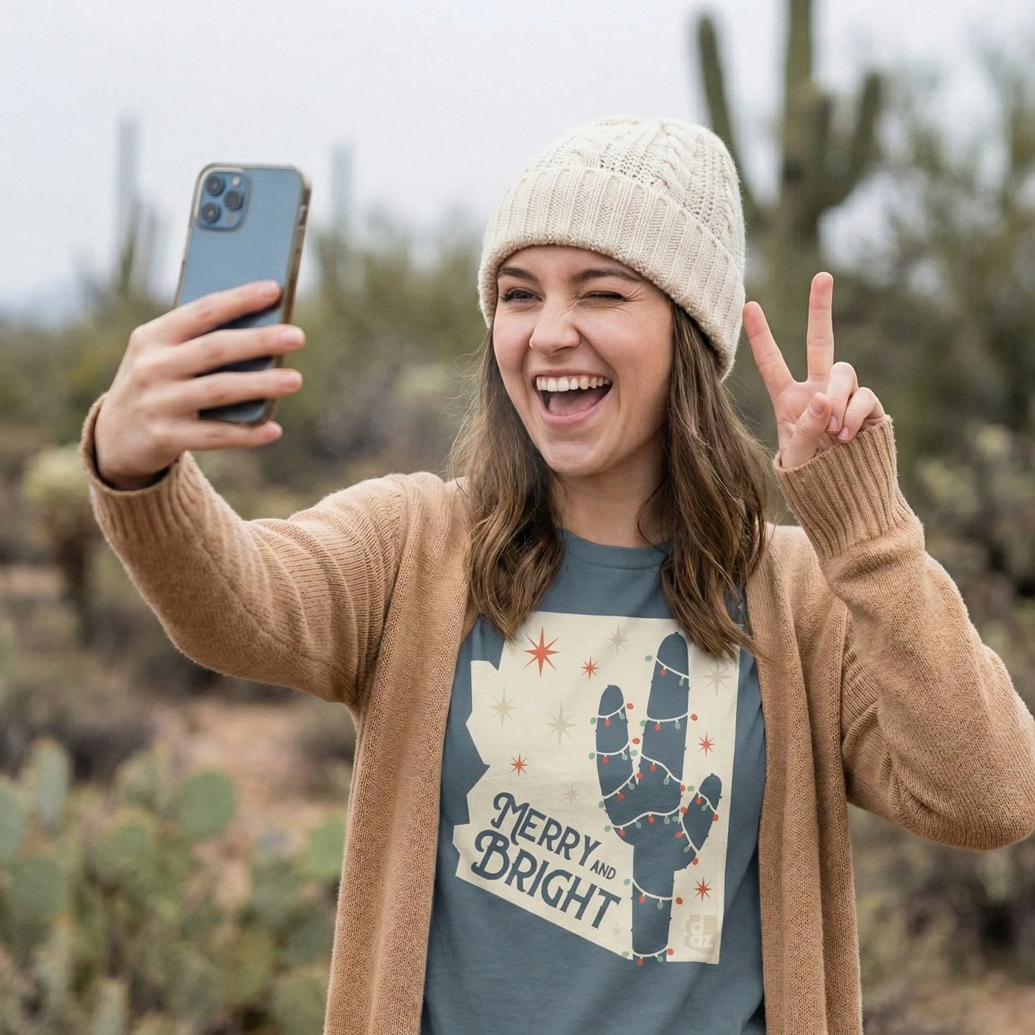 Woman wearing a beige beanie and brown cardigan taking a selfie outdoors, making a peace sign with her right hand, smiling and winking, with cacti in the background.