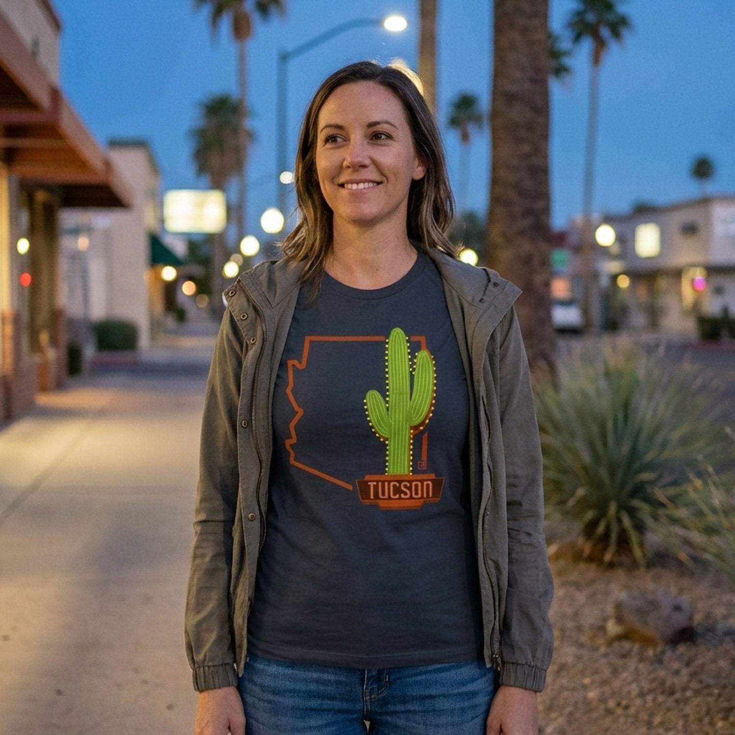 A woman standing outside at dusk on a street with palm trees in the background. She is wearing a dark t-shirt with a cactus graphic and the word 'Tucson' and a gray jacket, smiling and looking to the side.