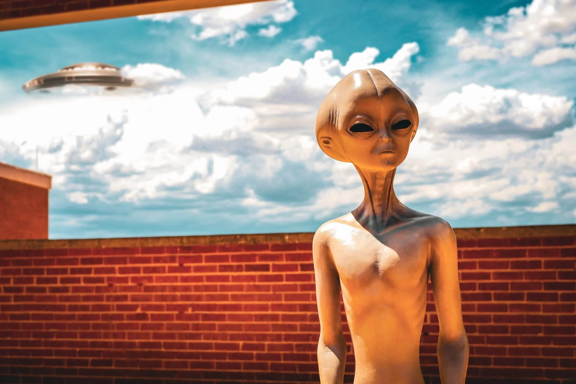 An alien figure with large black eyes and a bald head standing outdoors against a red brick wall, with a blue sky and clouds in the background, and a flying saucer-shaped UFO in the sky.