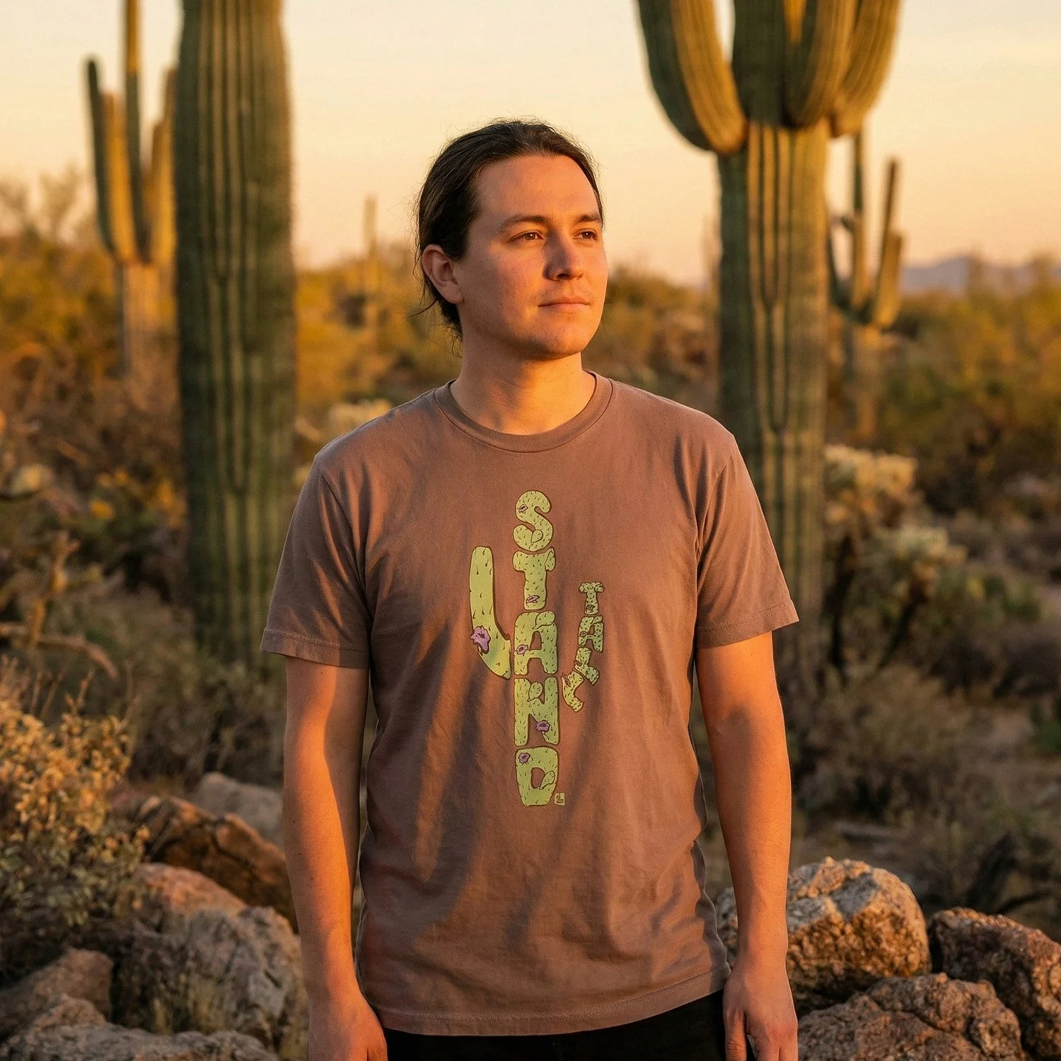 A young man with dark hair standing outdoors in a desert landscape during sunset, wearing a brown T-shirt with green and purple graphic design.