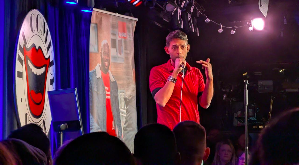 A male comedian, Kal Aise on stage performing at a comedy club called The Comedy Store with a microphone, audience in the foreground, club logo with laughing face on a blue curtain, and a poster promoting the charity Crisis in the background.