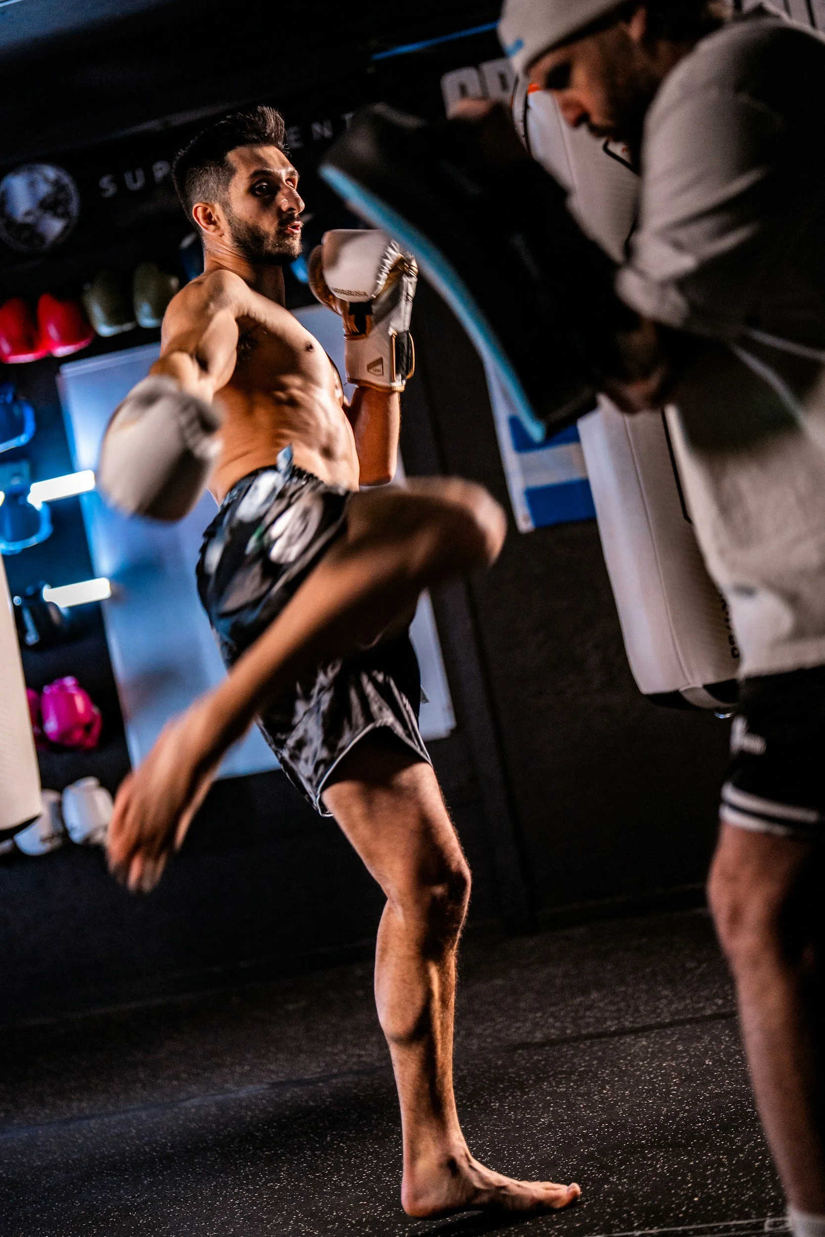 A shirtless male martial artist practicing kicks in a gym, wearing boxing gloves and shorts, with a trainer observing nearby.