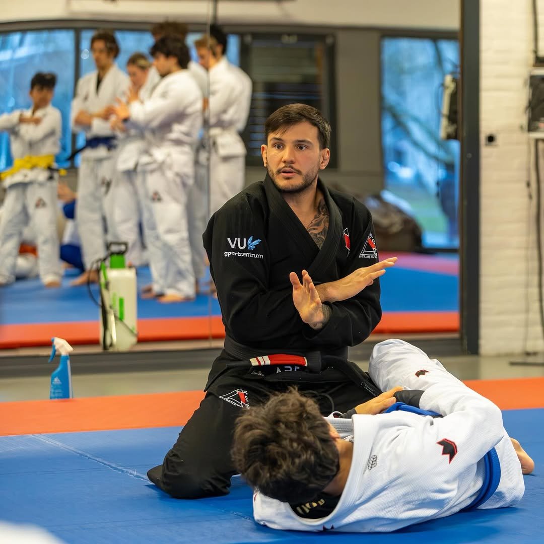Martial arts instructor in black gi teaches Brazilian jiu-jitsu to student in white gi, inside gym with other students watching.
