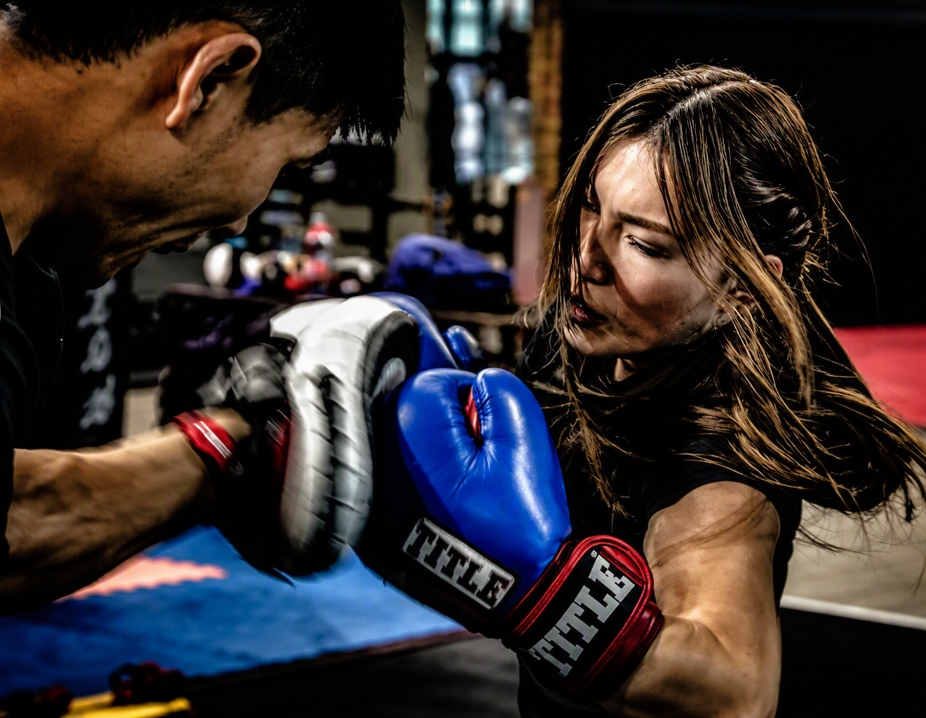 A woman practicing boxing with a man, both wearing boxing gloves, in a gym.