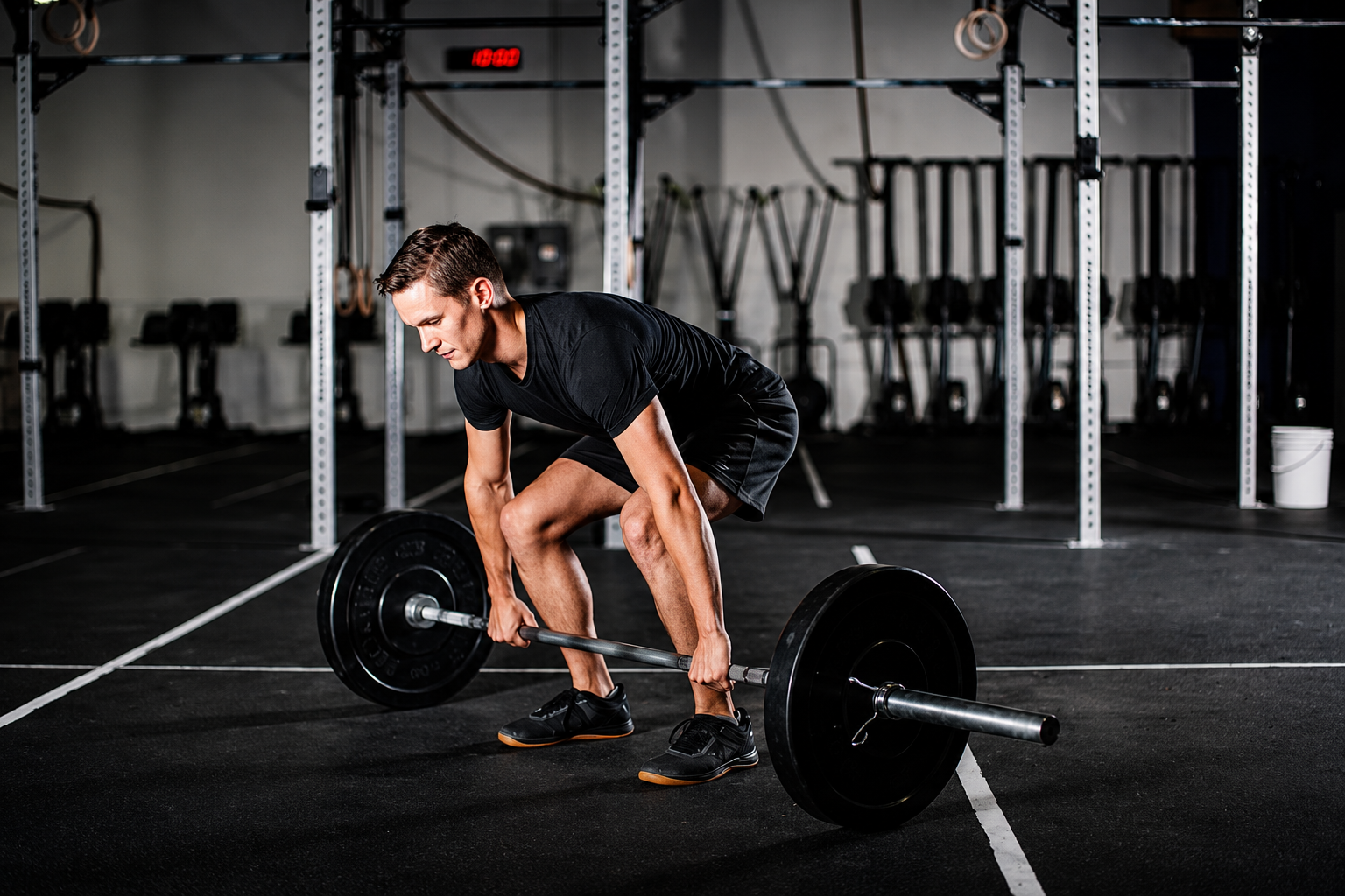A man in black athletic clothing lifting a barbell in a gym with black flooring, metal racks, and equipment in the background.