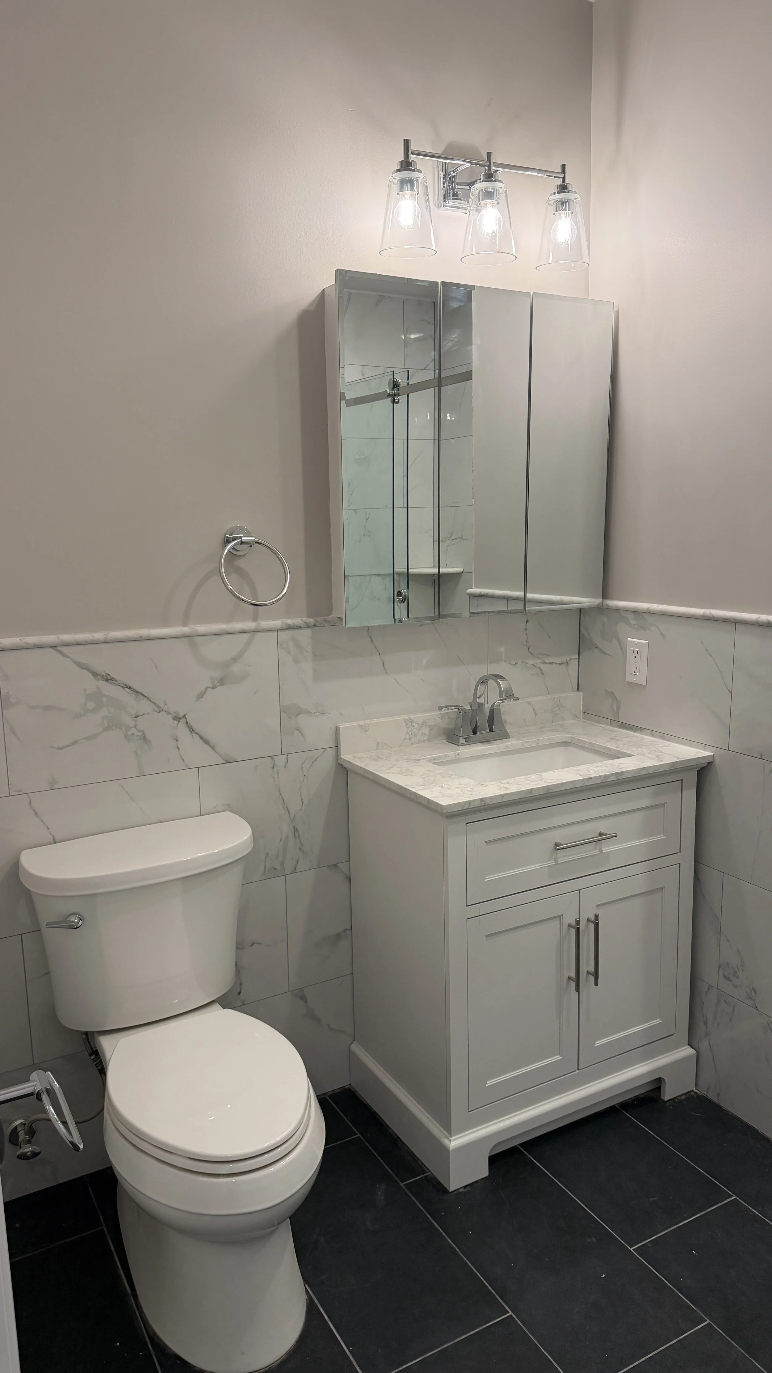 A modern bathroom with a white toilet, a white vanity with a marble countertop and a sink, a mirrored medicine cabinet, and a wall-mounted light fixture with three glass shades.