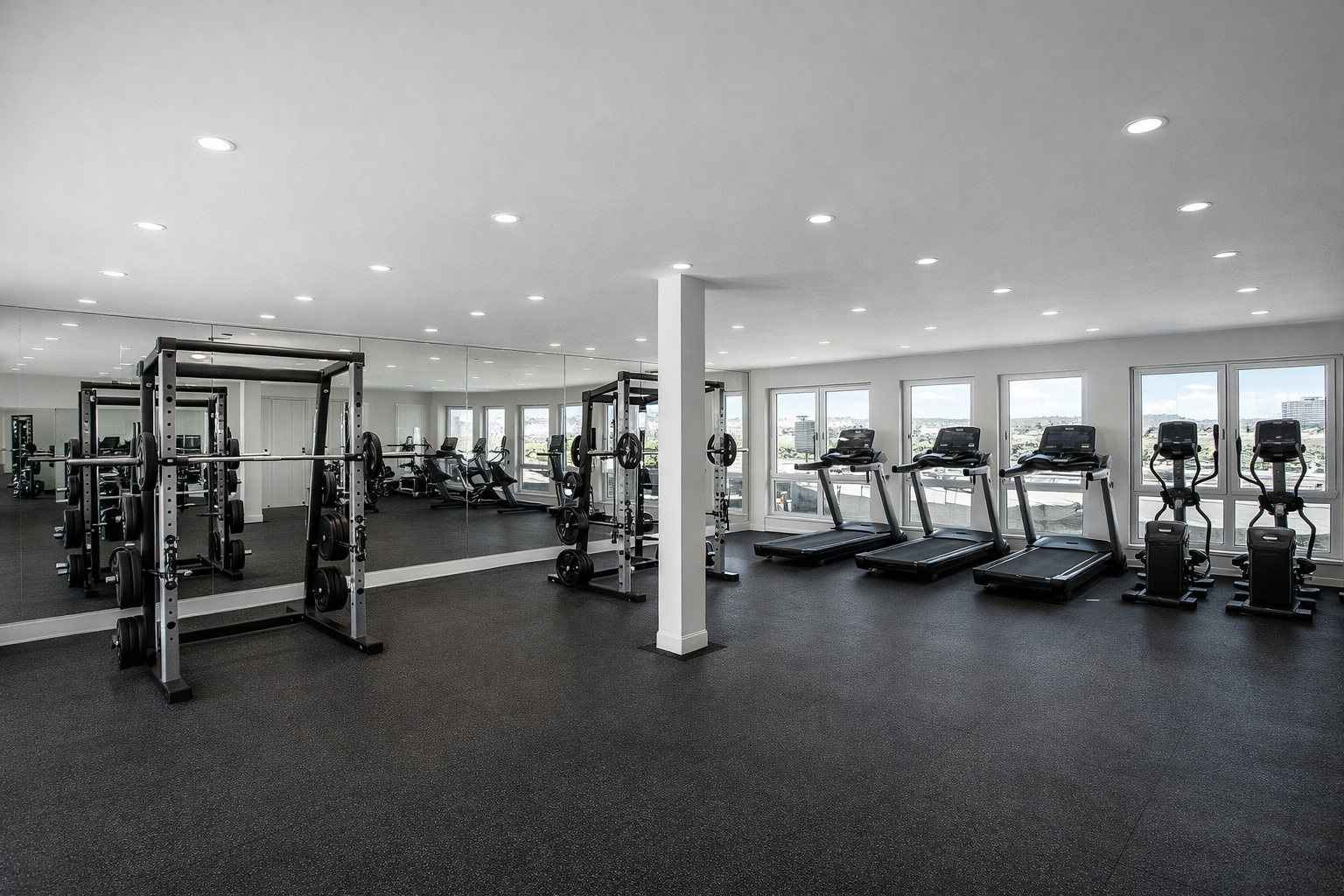 Empty gym room with weightlifting equipment, treadmills, and stationary bikes, large windows showing city view, bright lighting.