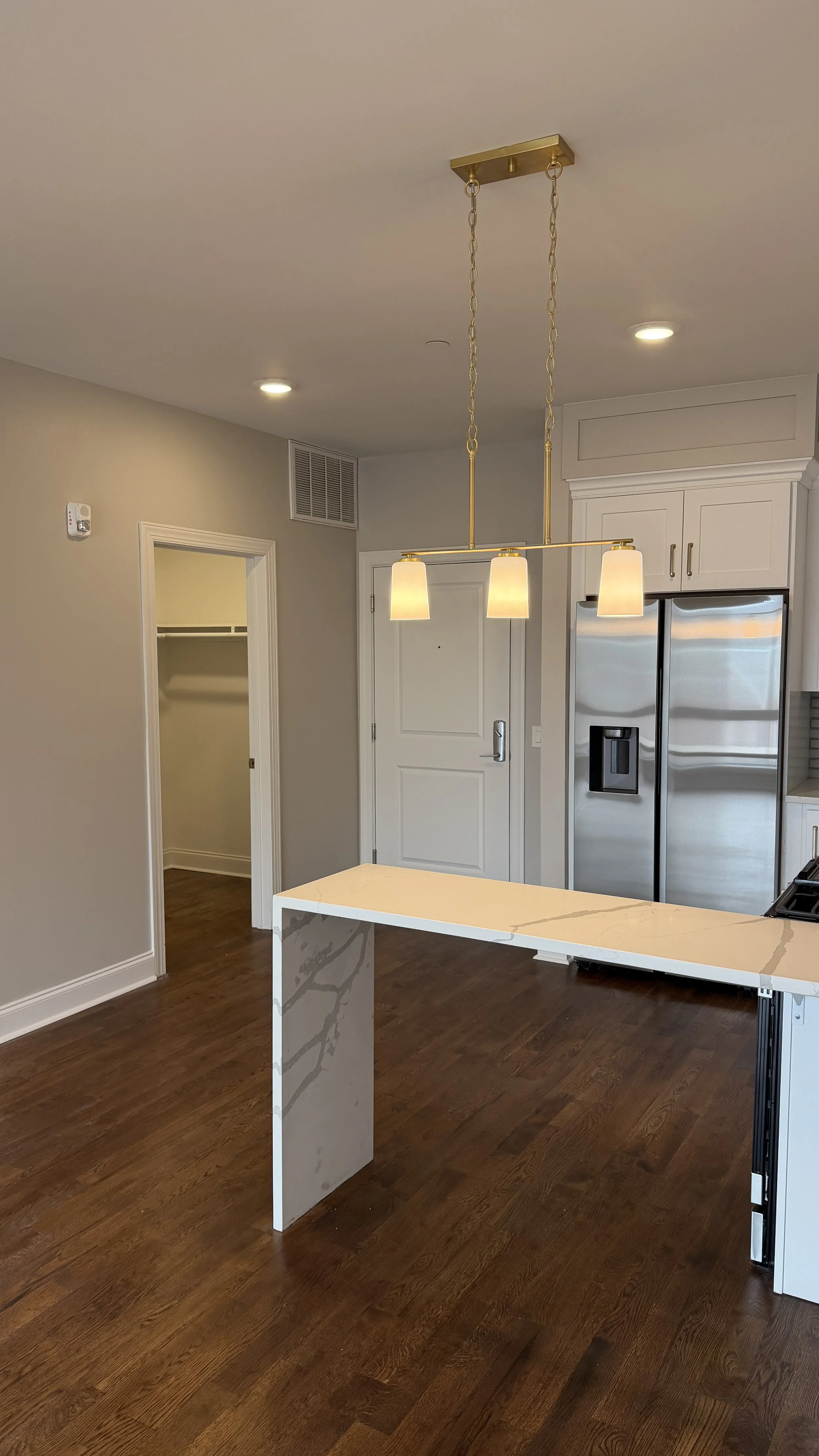 Modern kitchen with a white marble countertop island, stainless steel refrigerator, white cabinets, and a gold hanging light fixture. There is an open closet door in the background.