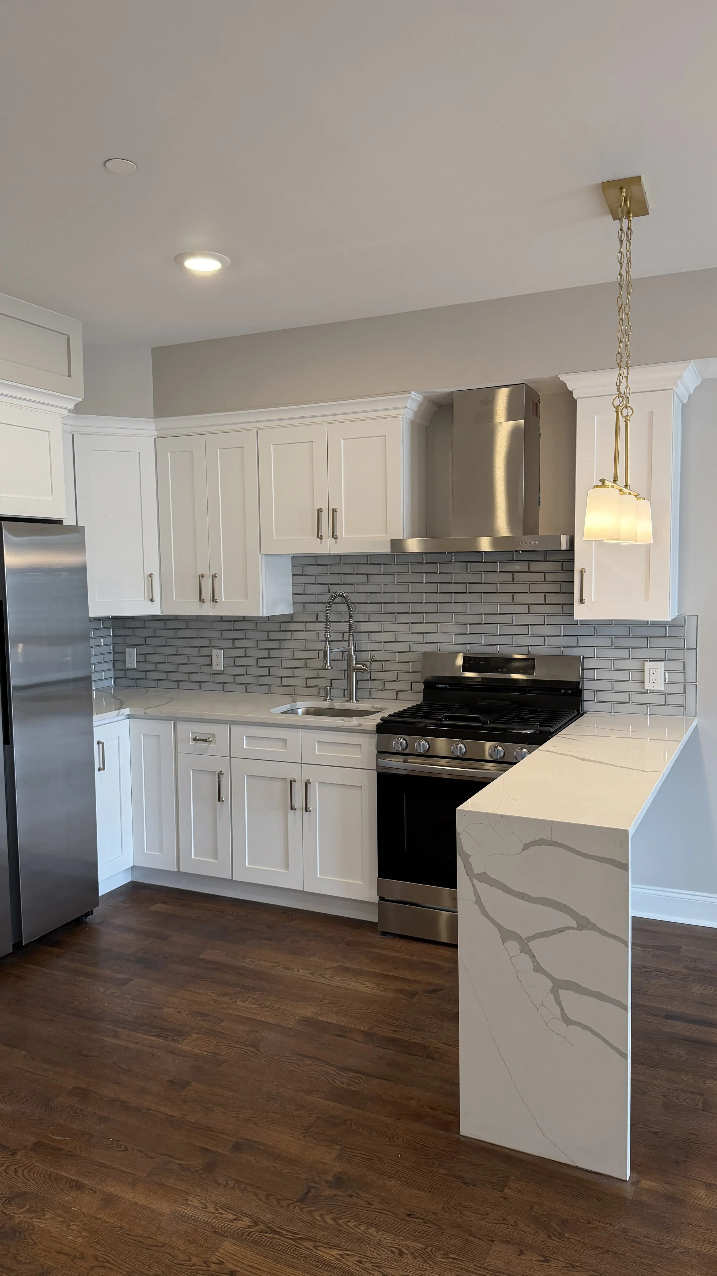 Modern kitchen with white cabinets, stainless steel appliances, gray tile backsplash, wooden floor, marble countertop with gray veining, and a pendant light.