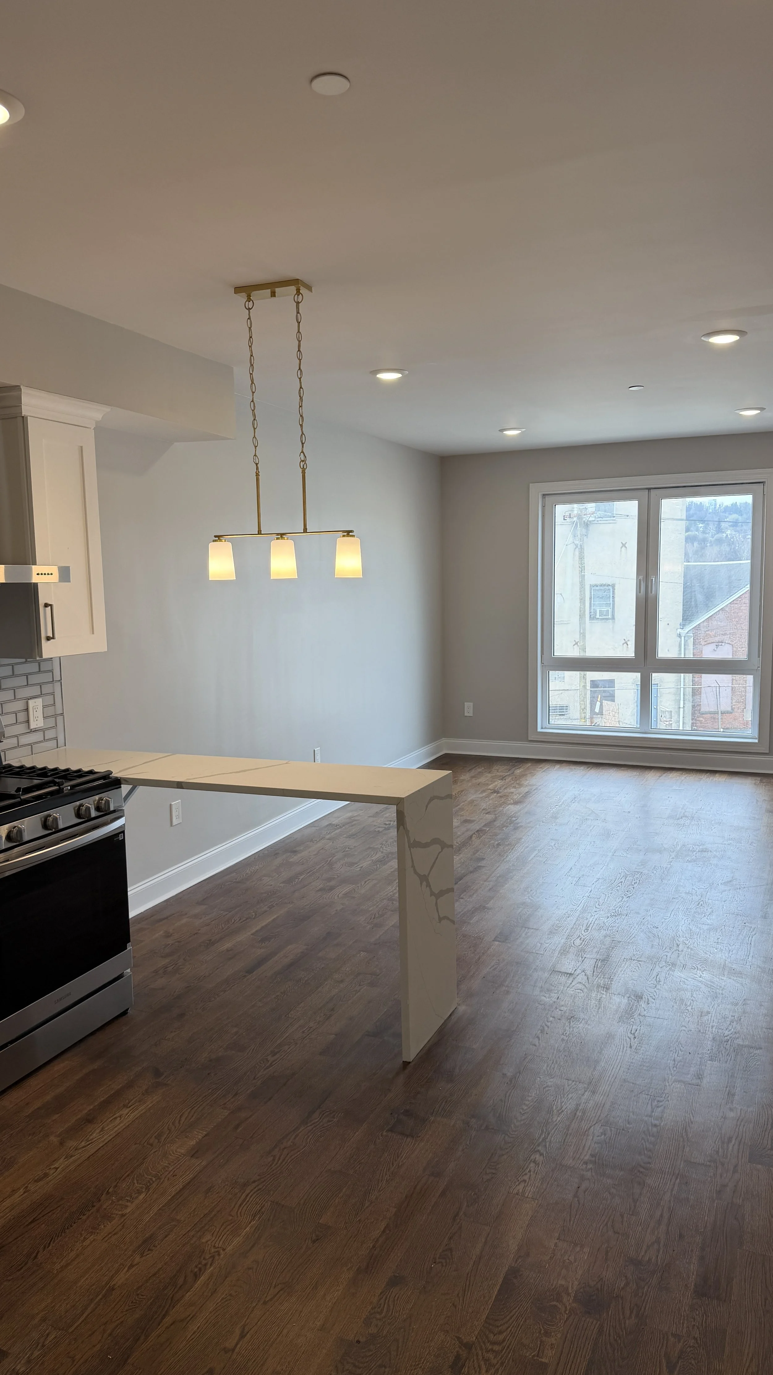 Empty living room and kitchen with hardwood floors, white cabinets, a window, and a modern pendant light fixture.