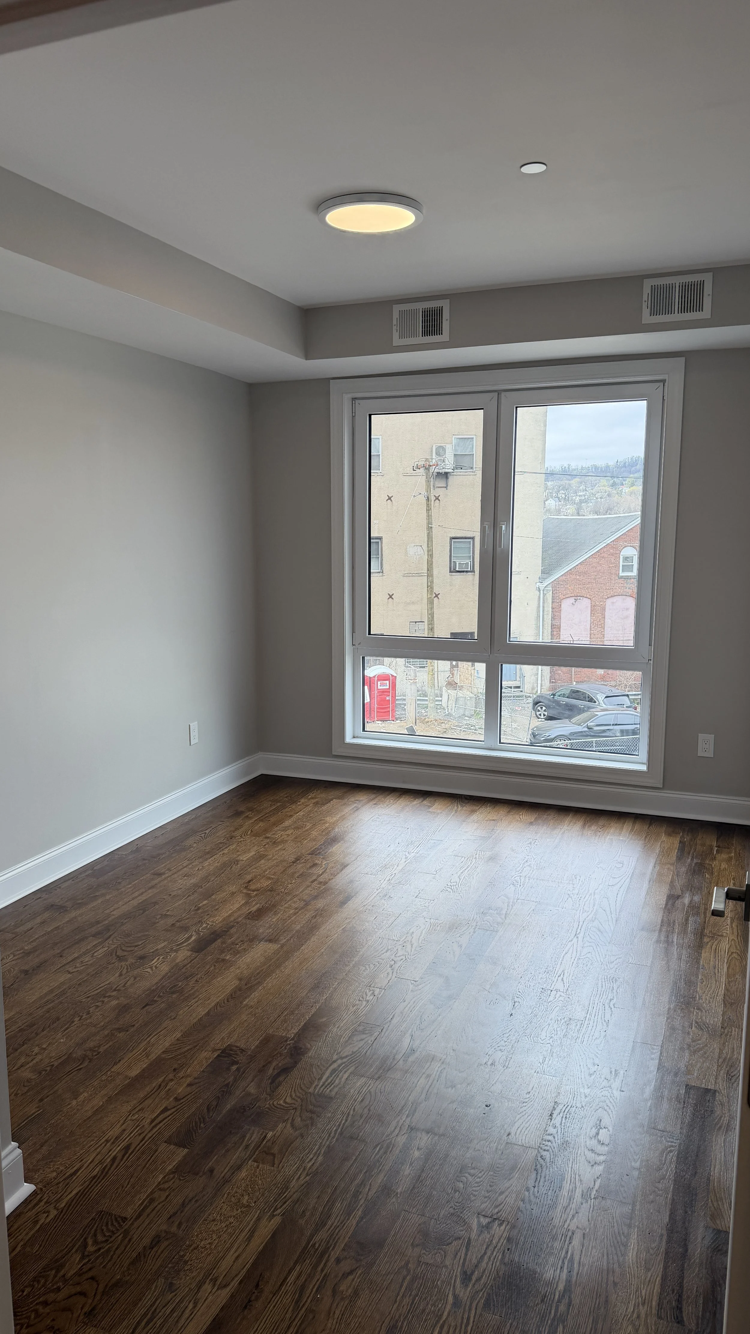 Empty room with wood flooring and large window showing a building and parked cars outside.